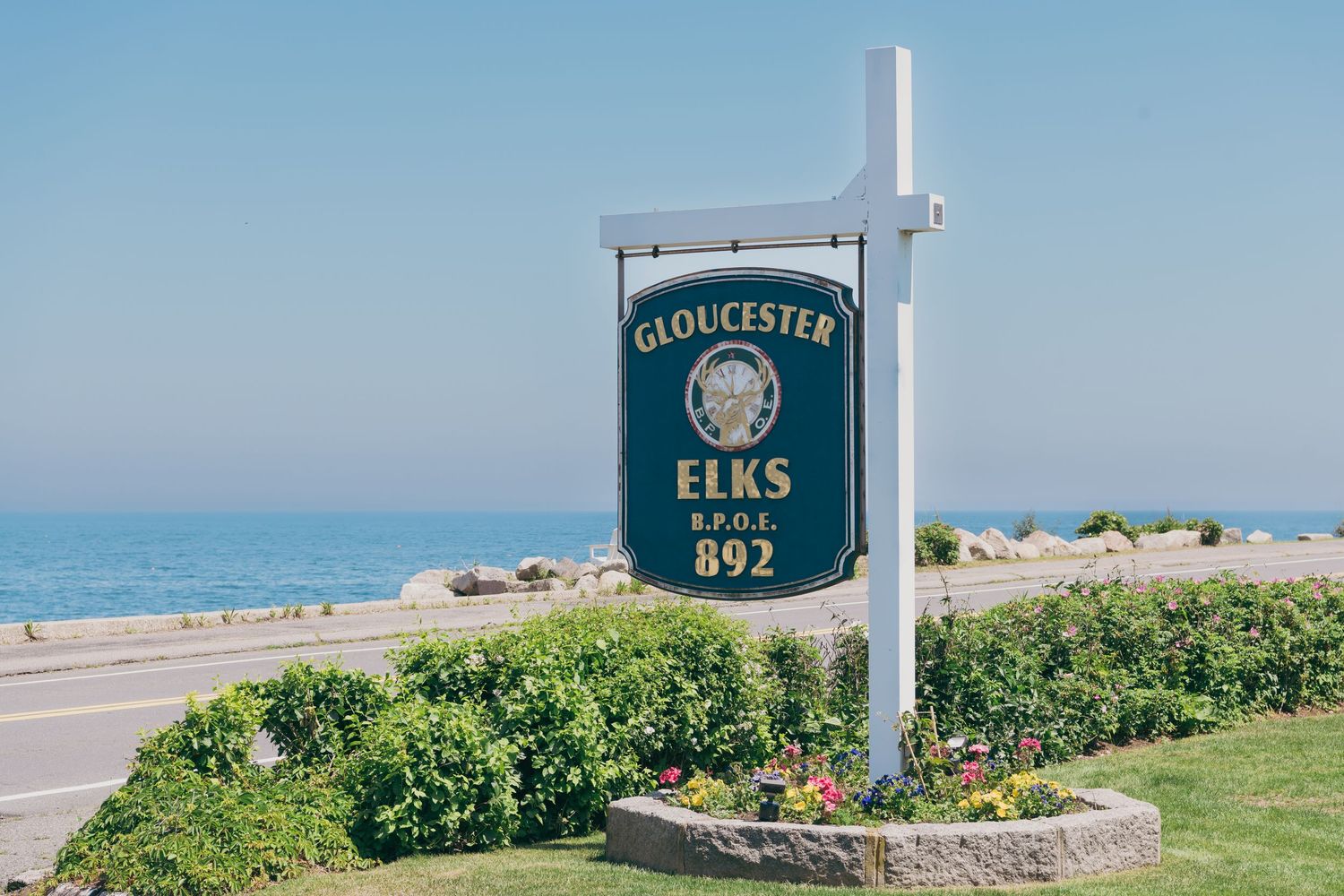 A scenic row of Gloucester Elks signs and markers line a coastal path with blue ocean views and green landscaping.