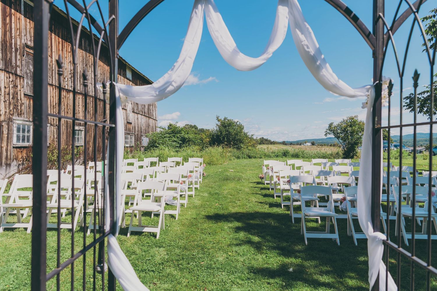 White ribbons and decor adorn metal gates in an outdoor wedding ceremony setup with white chairs on grassy lawn.