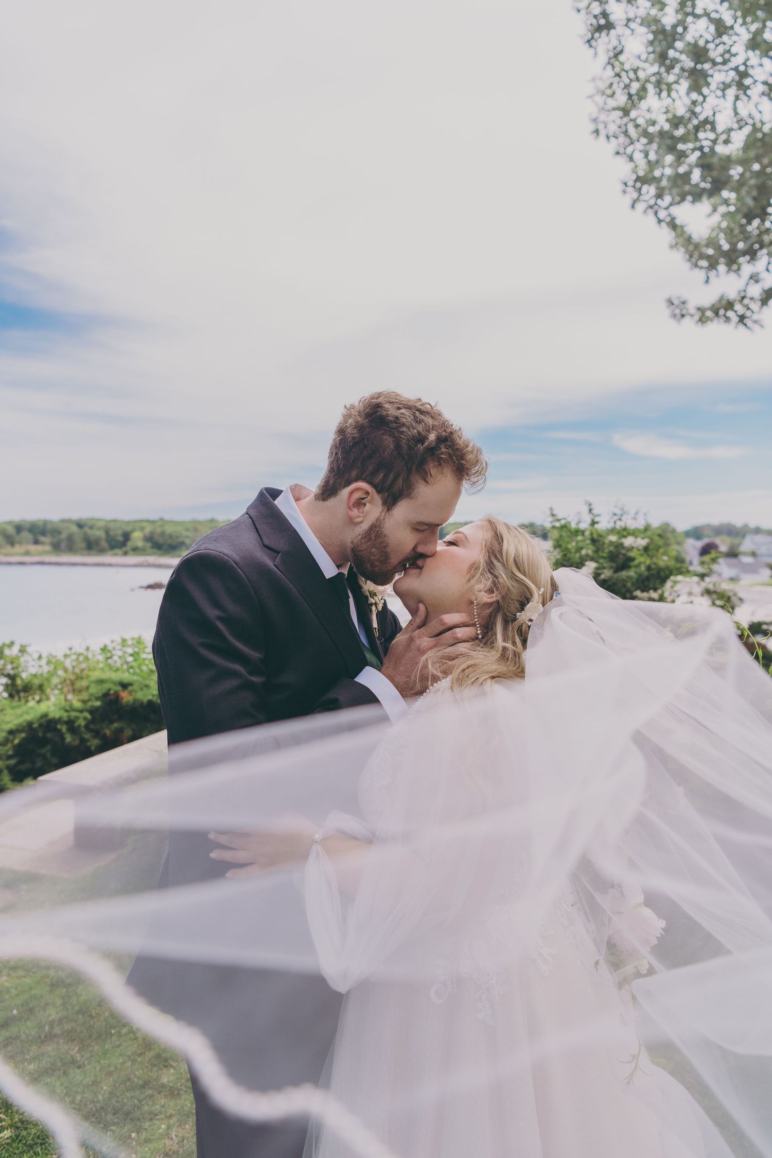A romantic wedding couple share a tender kiss outdoors with a flowing veil and waterfront backdrop.