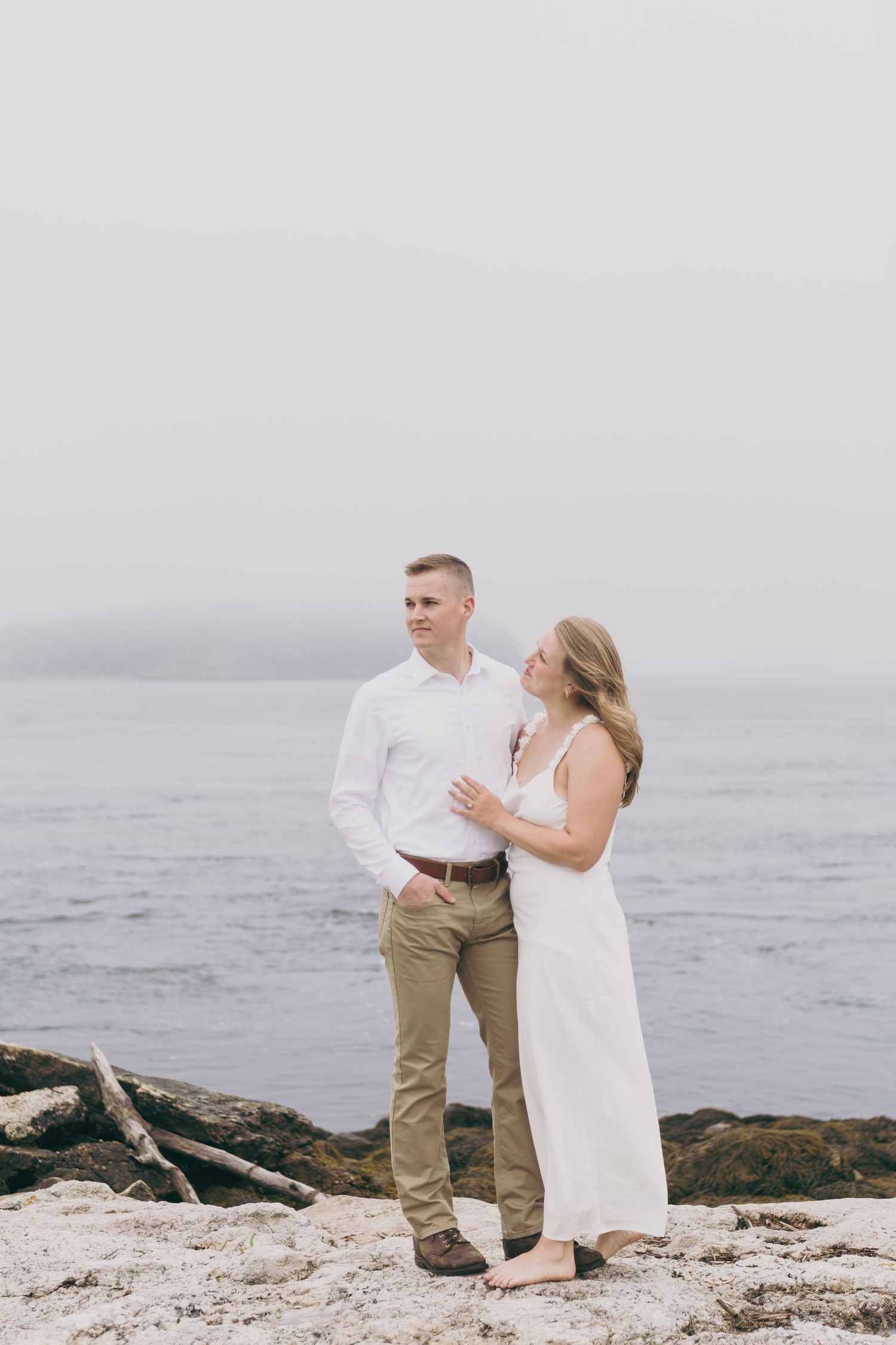 A couple in casual attire shares a romantic moment on a misty rocky coastline at the beach.