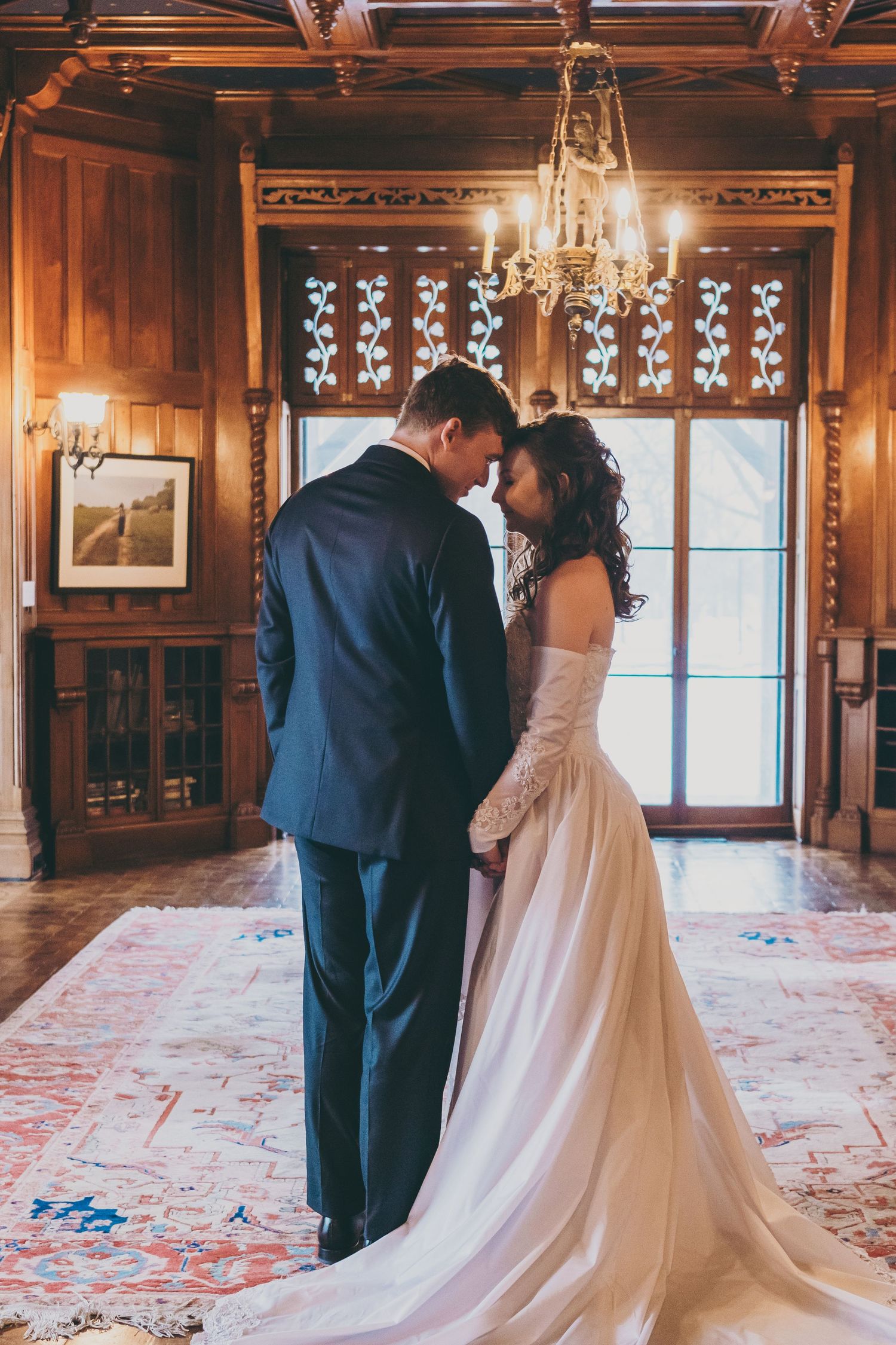A romantic wedding moment in an elegant wood-paneled room with a chandelier and ornate window.