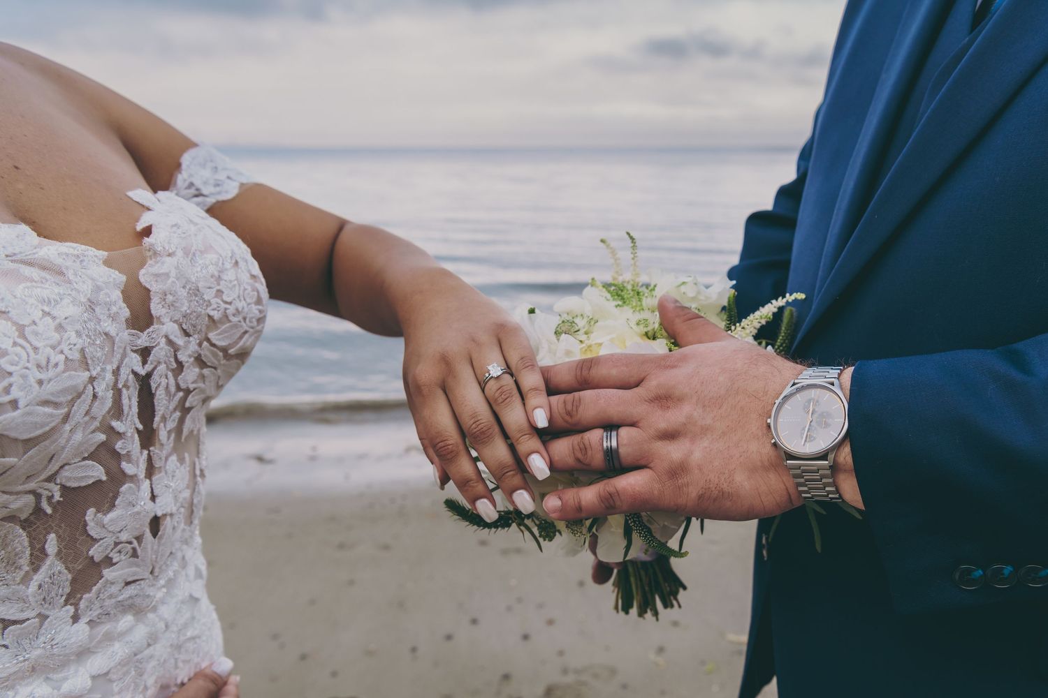 Wedding couple holding hands with elegant bouquet on a cloudy beach with waves in the background.