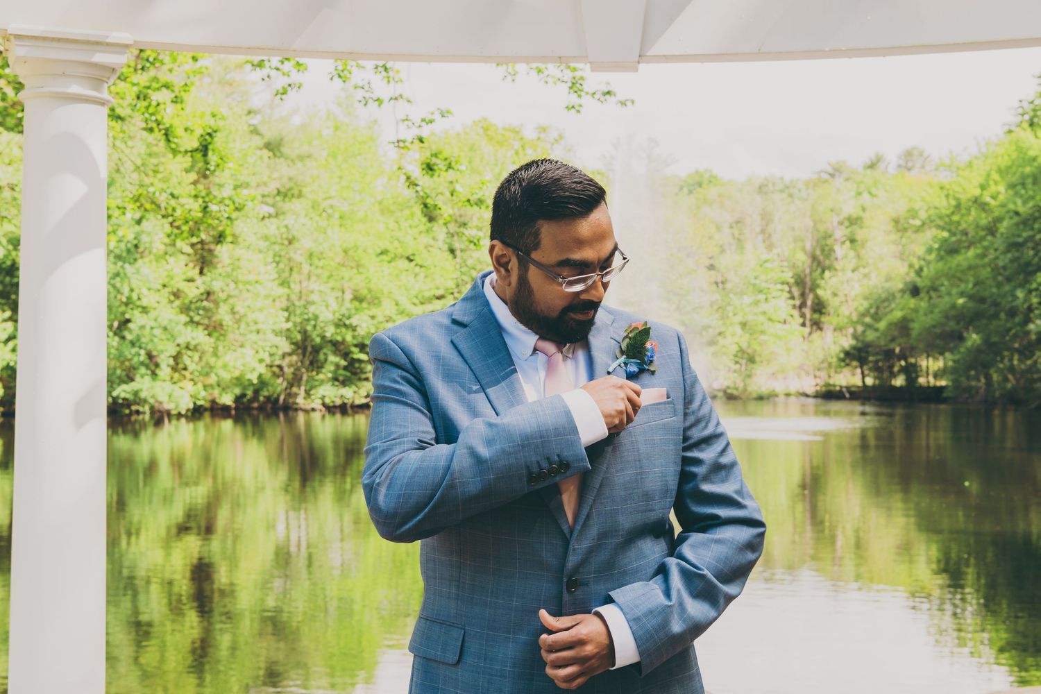 A groom in a light blue suit adjusts his lapel while standing by a tranquil pond surrounded by spring greenery.