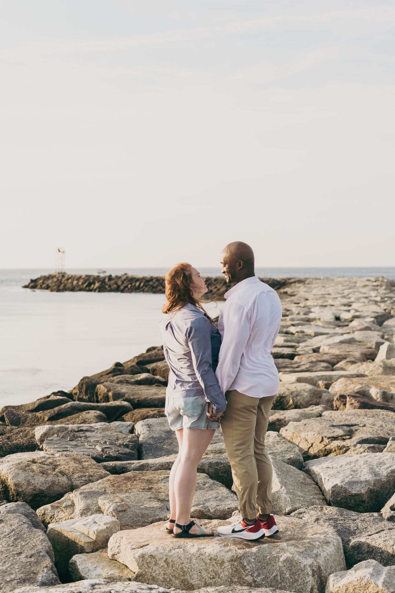 A couple stands together on rocky coastal breakwater during sunset by the ocean.
