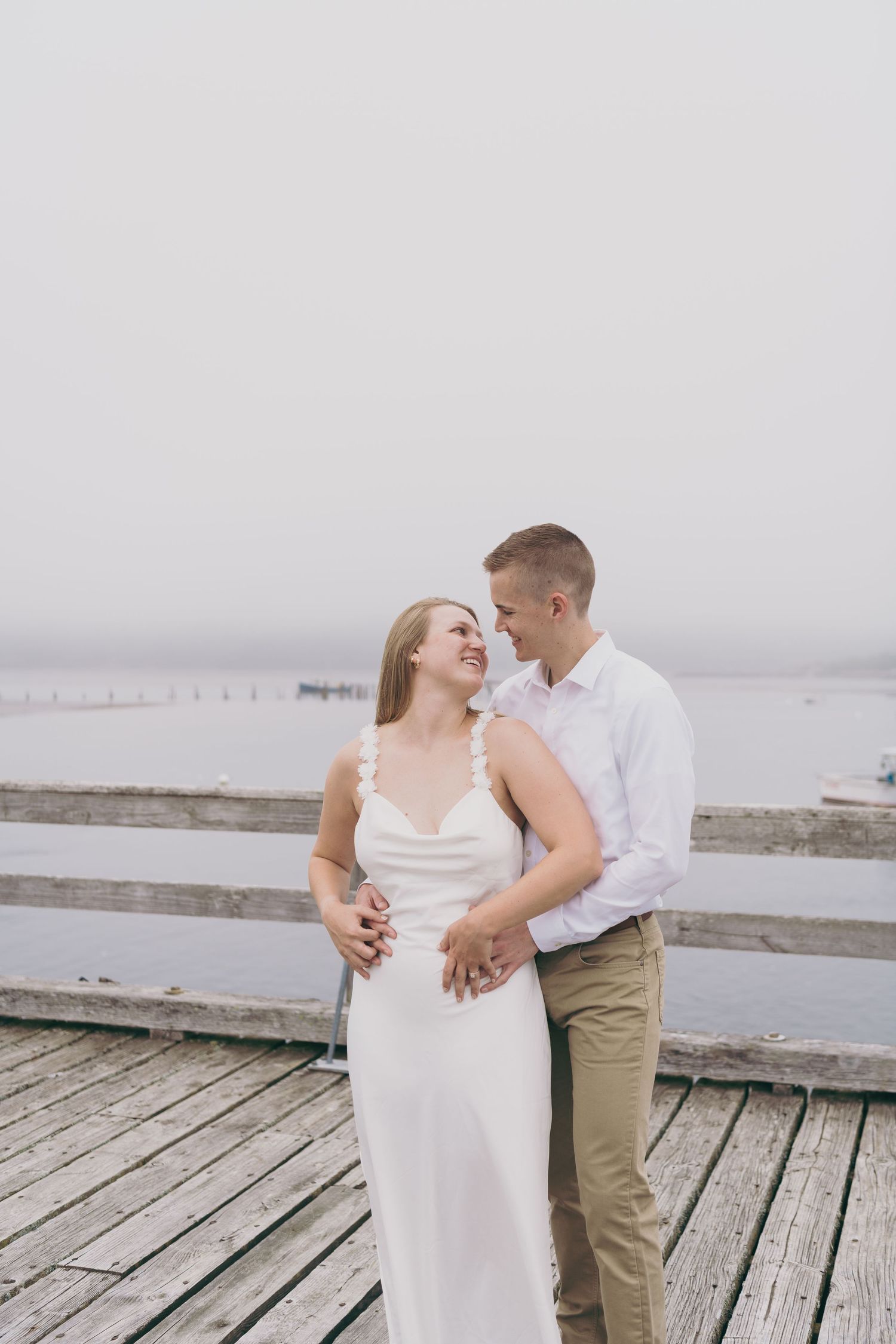 A romantic couple shares a tender moment on a wooden dock overlooking a misty waterfront.