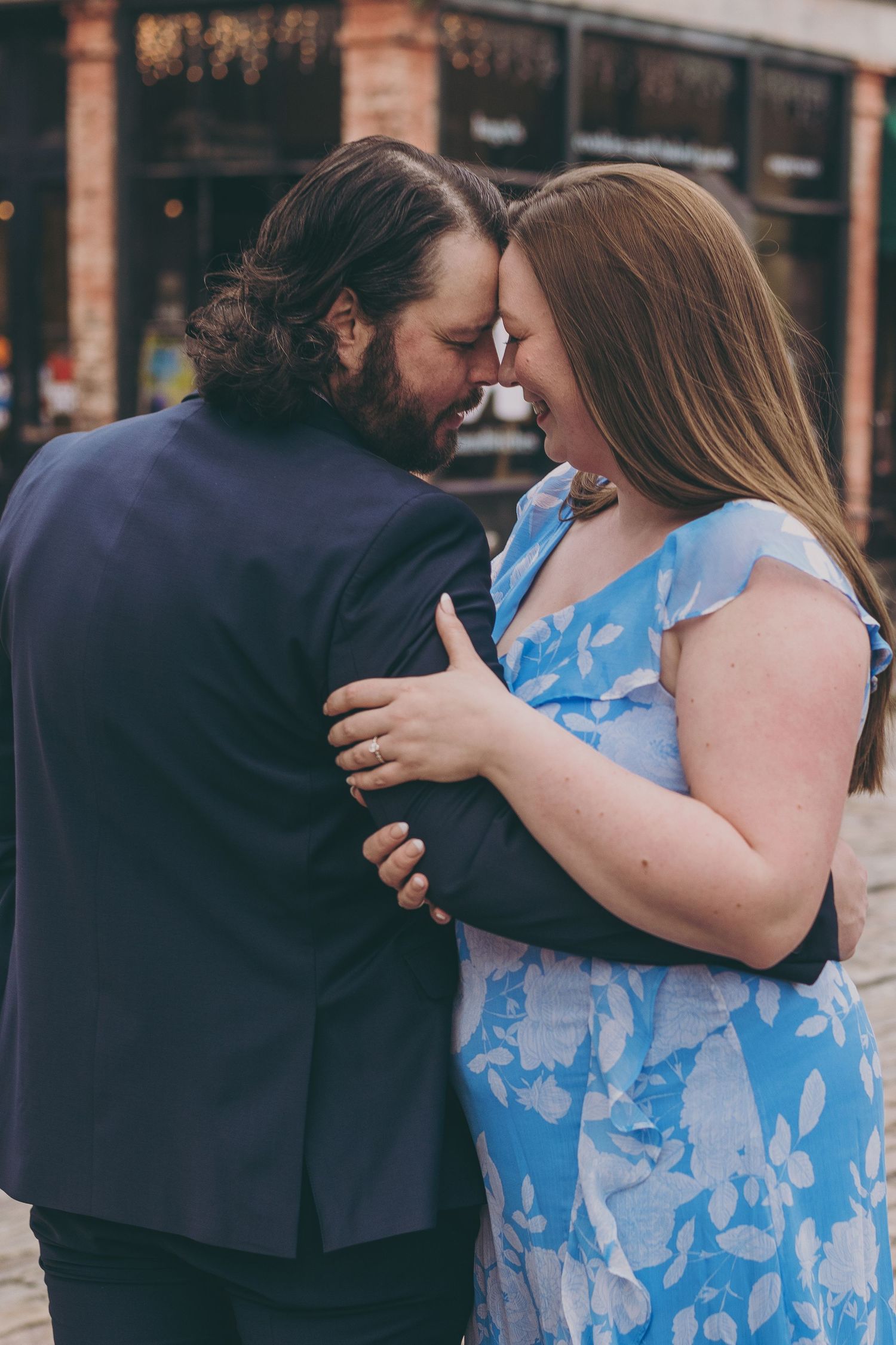 A couple embraces outdoors, one wearing a blue floral dress and the other in a dark suit jacket.