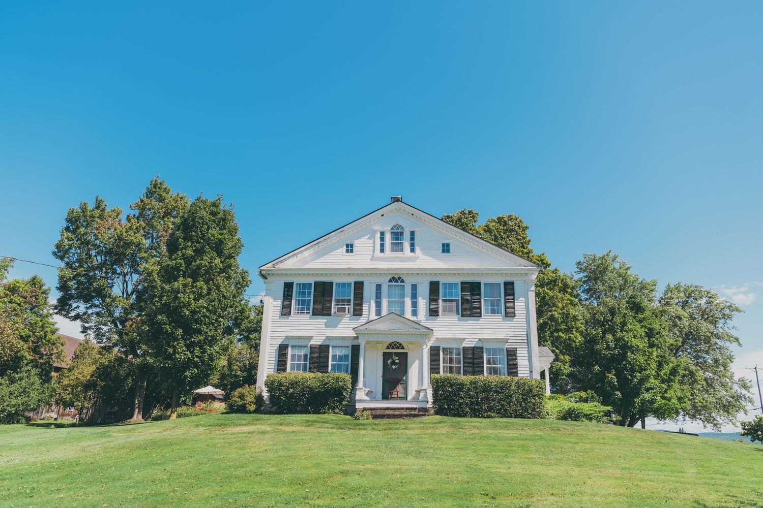 White colonial style house on grassy hill with blue sky and trees in panoramic landscape view.