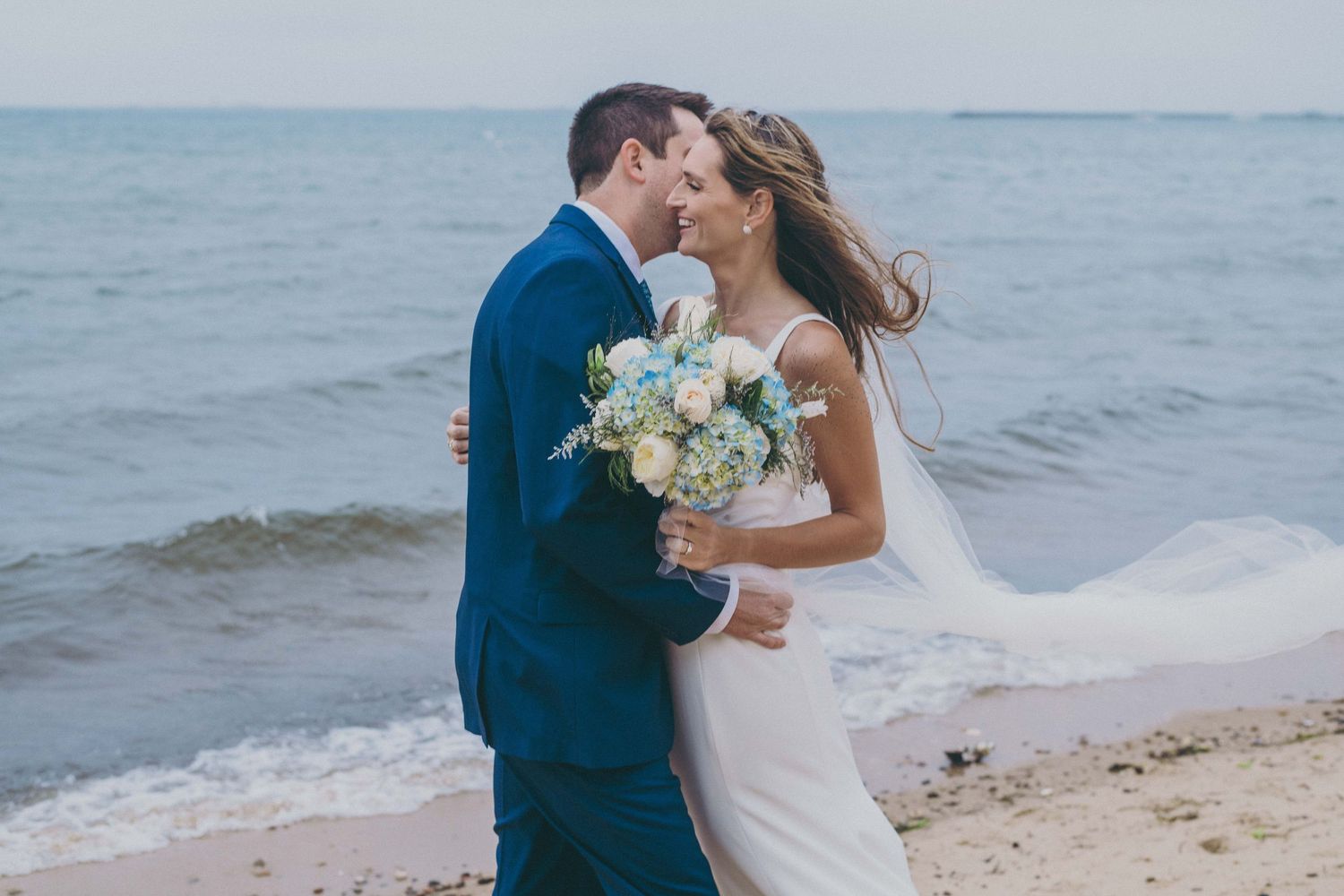 Couple in wedding attire embracing on a beach with ocean waves in the background.