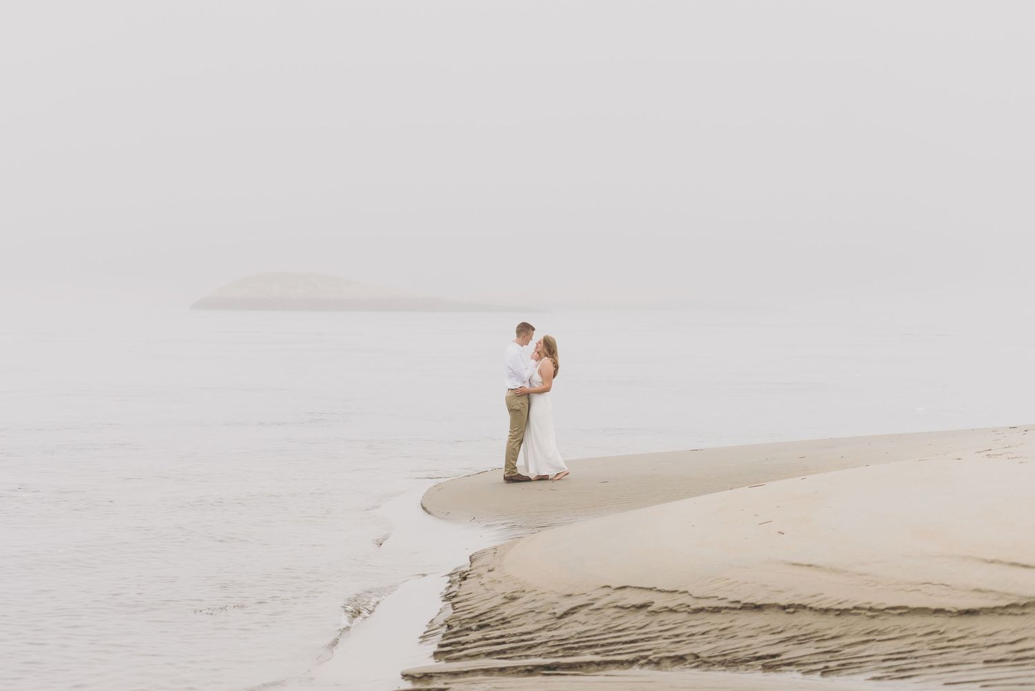 A couple embraces on a misty sand dune overlooking a foggy beach landscape.