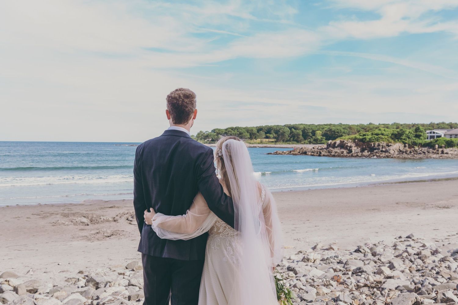 A couple walks along a scenic beach shore with homes and palm trees visible in the distant coastline.