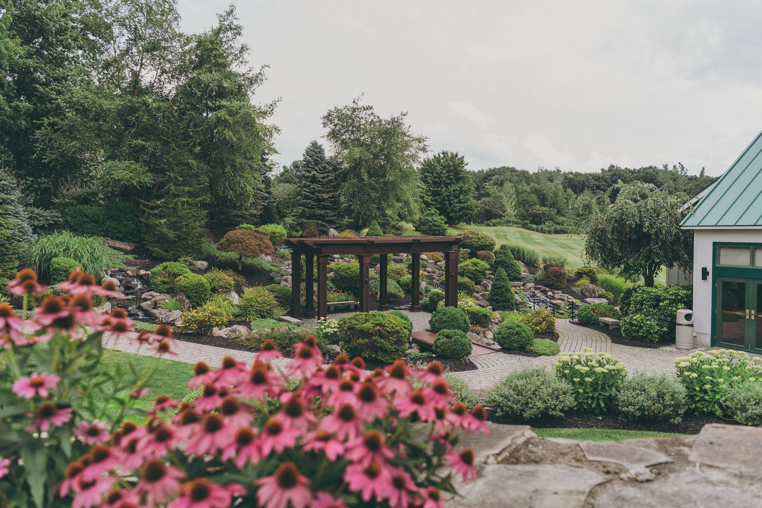 A beautiful landscaped garden with pink flowers and a wooden pergola structure surrounded by lush greenery.