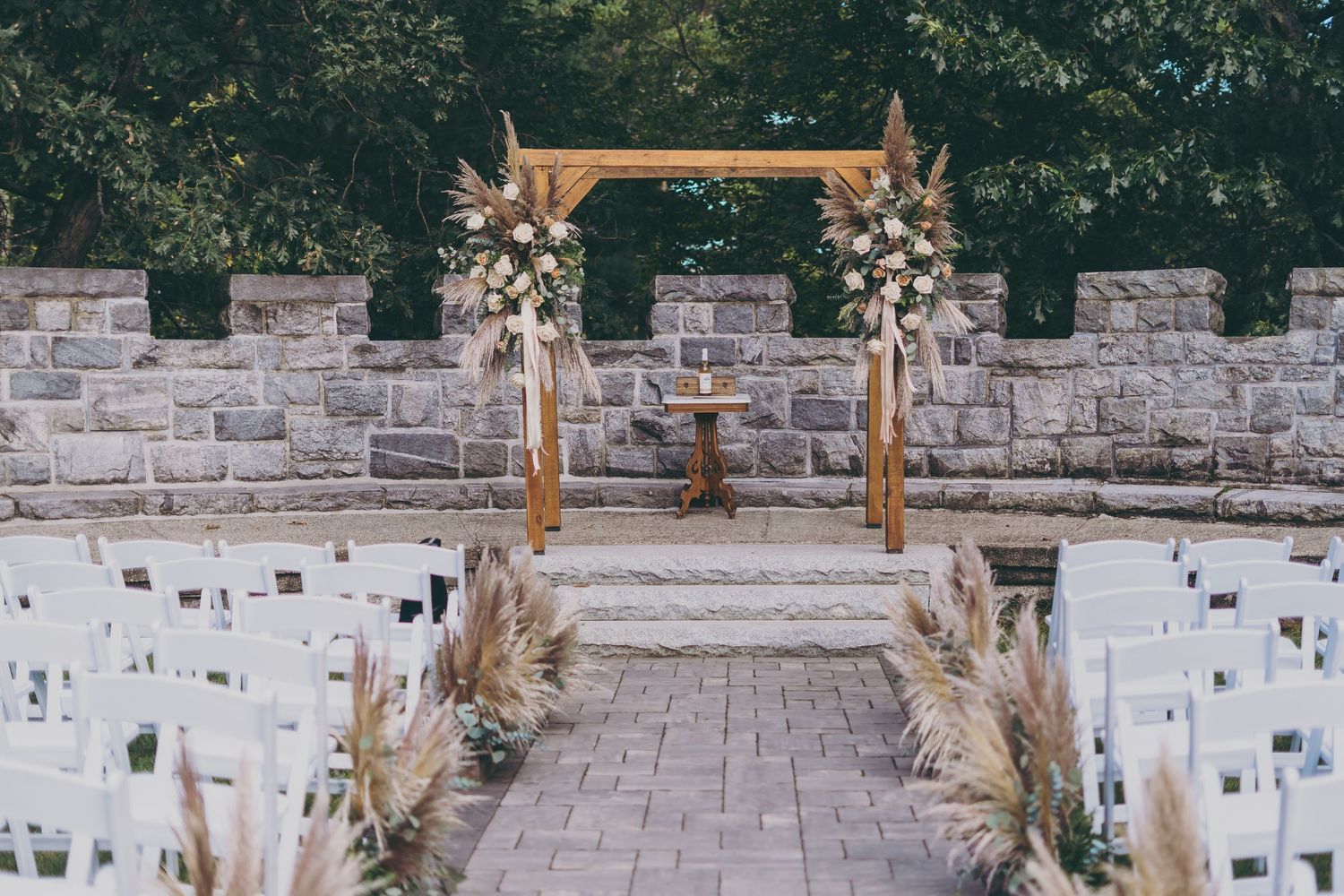 Rustic wooden wedding arch decorated with pampas grass and white florals against a stone wall with white chairs lined up.