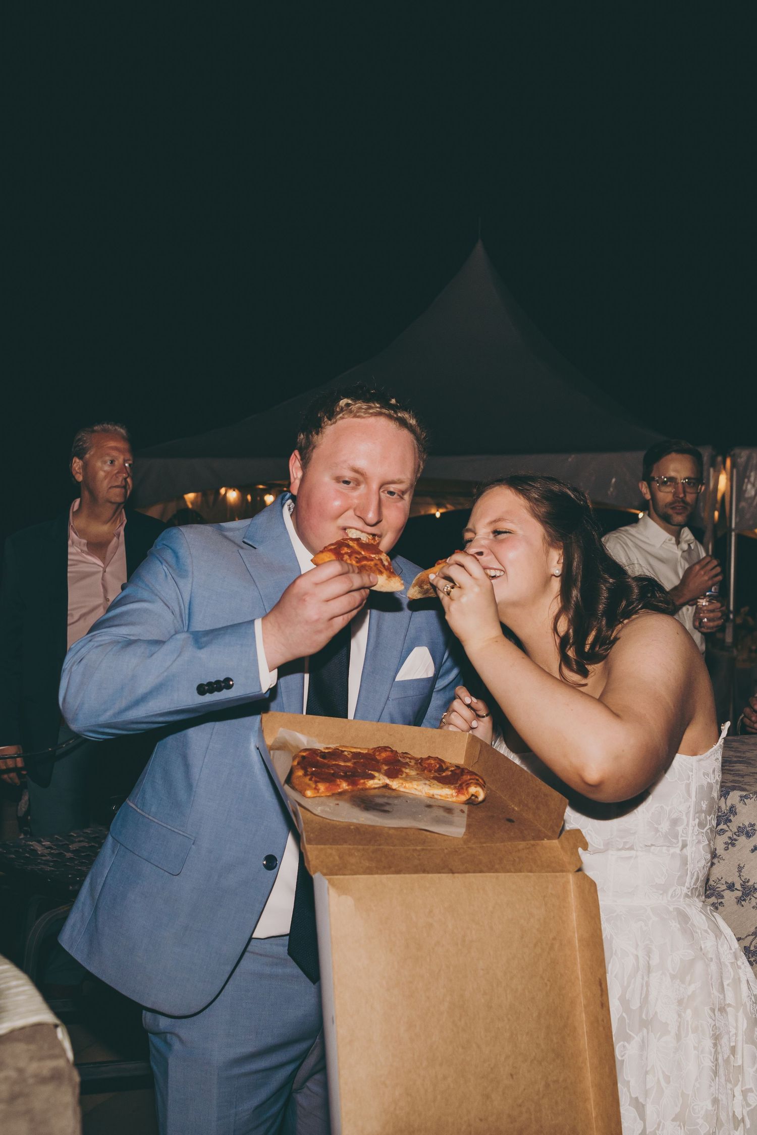 Couple sharing pizza from a box during their wedding reception at night.