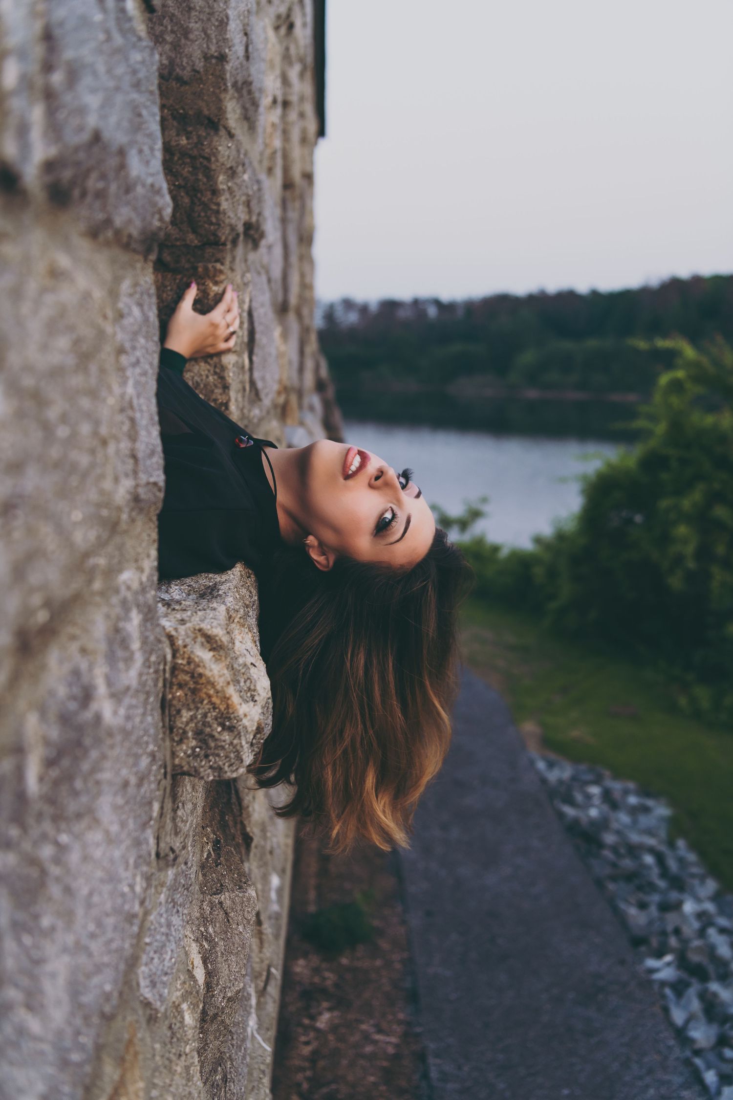 Artistic portrait against a stone wall with lakeside scenery in the background.