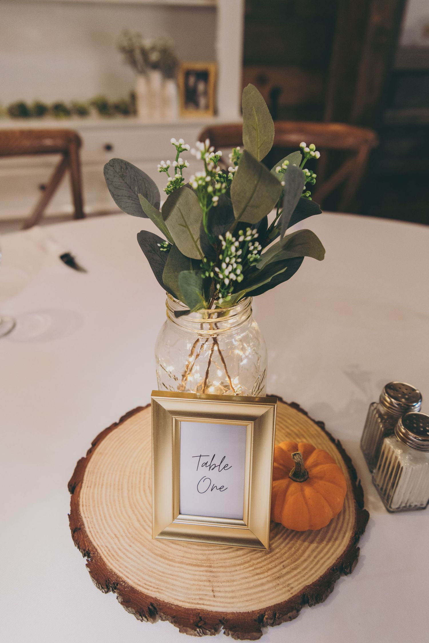 Wedding table centerpiece with eucalyptus in mason jar, wood slice, pumpkin, and 'Table One' frame.