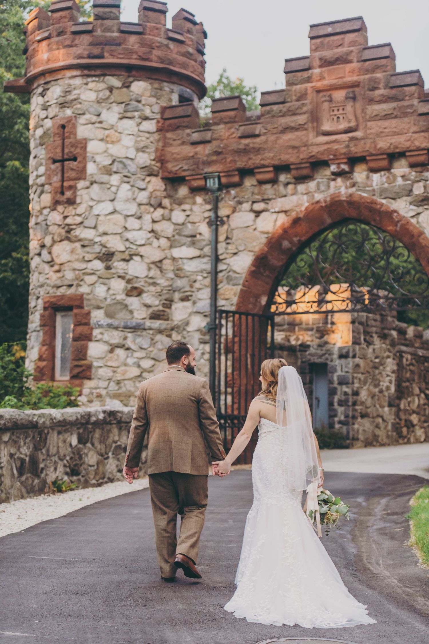 A couple in wedding attire walk hand in hand towards a beautiful stone castle entrance with crenellated towers.