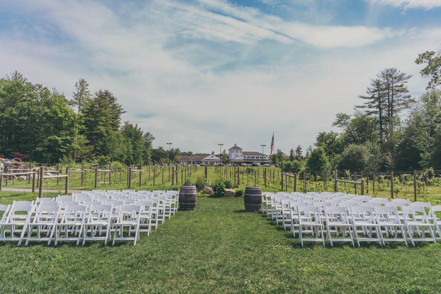 White wedding chairs arranged in rows on a green lawn with vineyard and trees in the background.