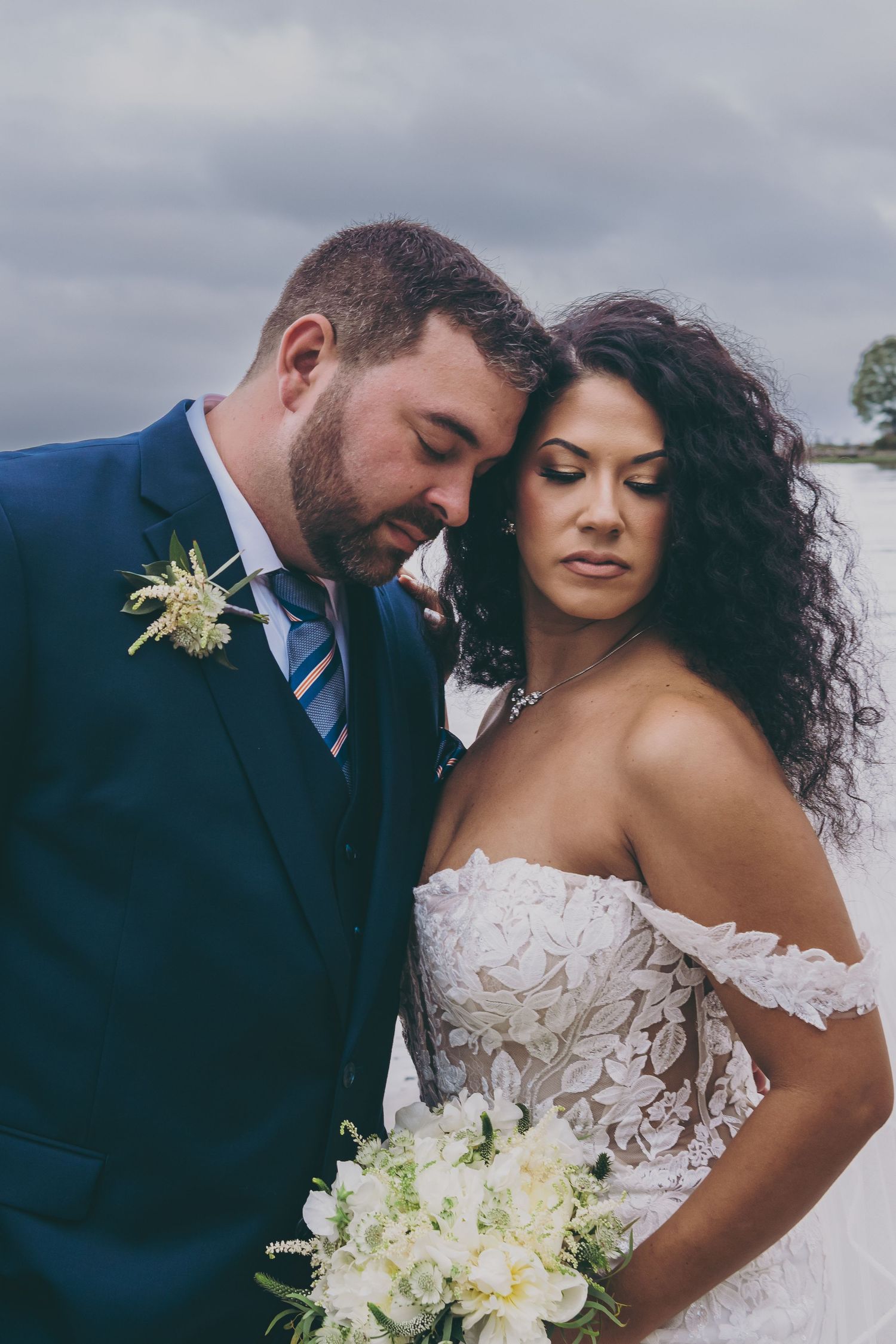 A couple shares an intimate moment in formal wedding attire, with the bride holding a white bouquet against a cloudy sky.