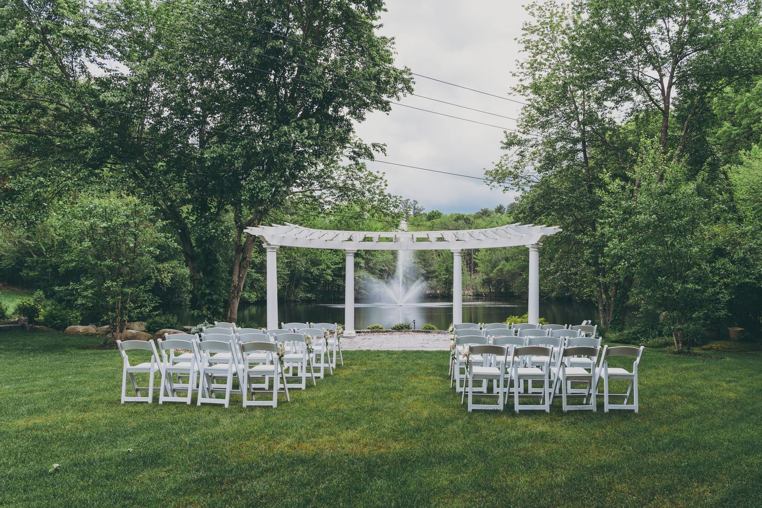 White wedding ceremony setup with pergola and folding chairs arranged on a lush green lawn surrounded by trees.