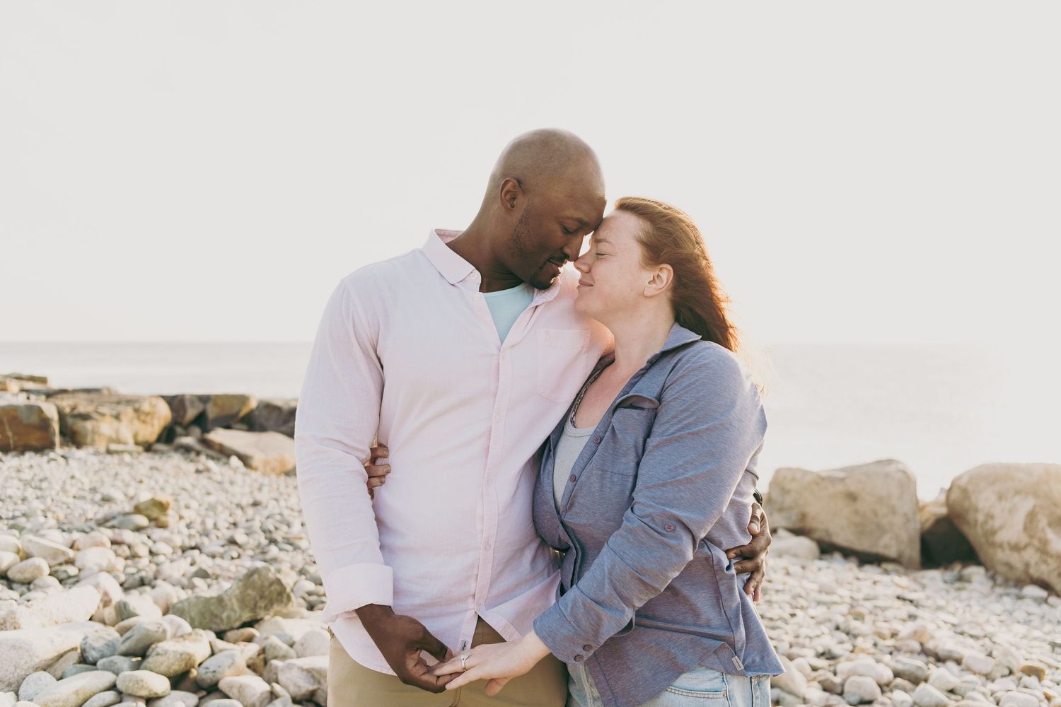 Romantic couple in casual attire embraces during sunset beach photoshoot at a rocky shoreline.