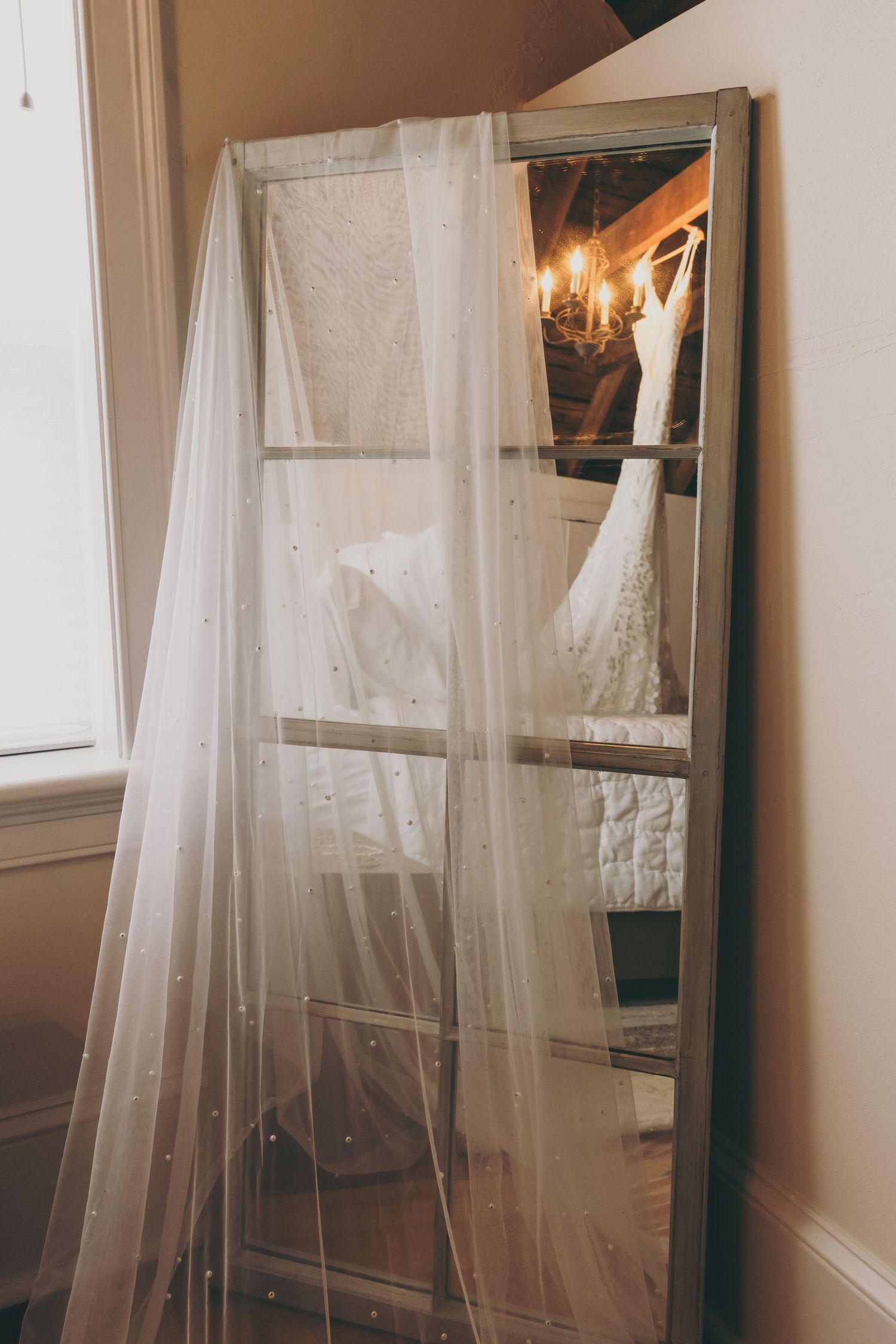 Wedding dress hanging on mirror with sheer white veil draped in soft natural light.