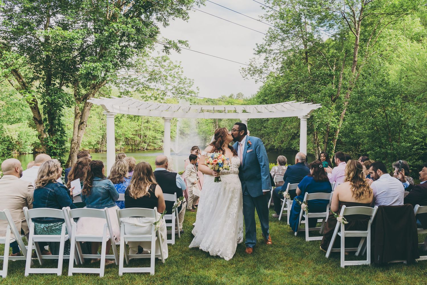 Outdoor wedding ceremony under white pergola with guests seated on folding chairs in garden setting.