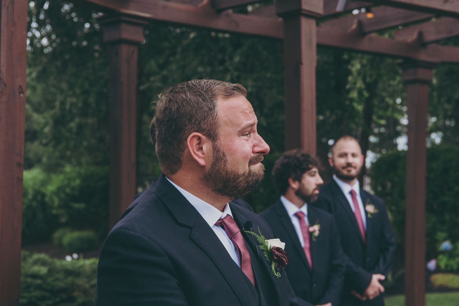 Wedding party members in black suits and red ties stand under a wooden pergola during an outdoor ceremony.
