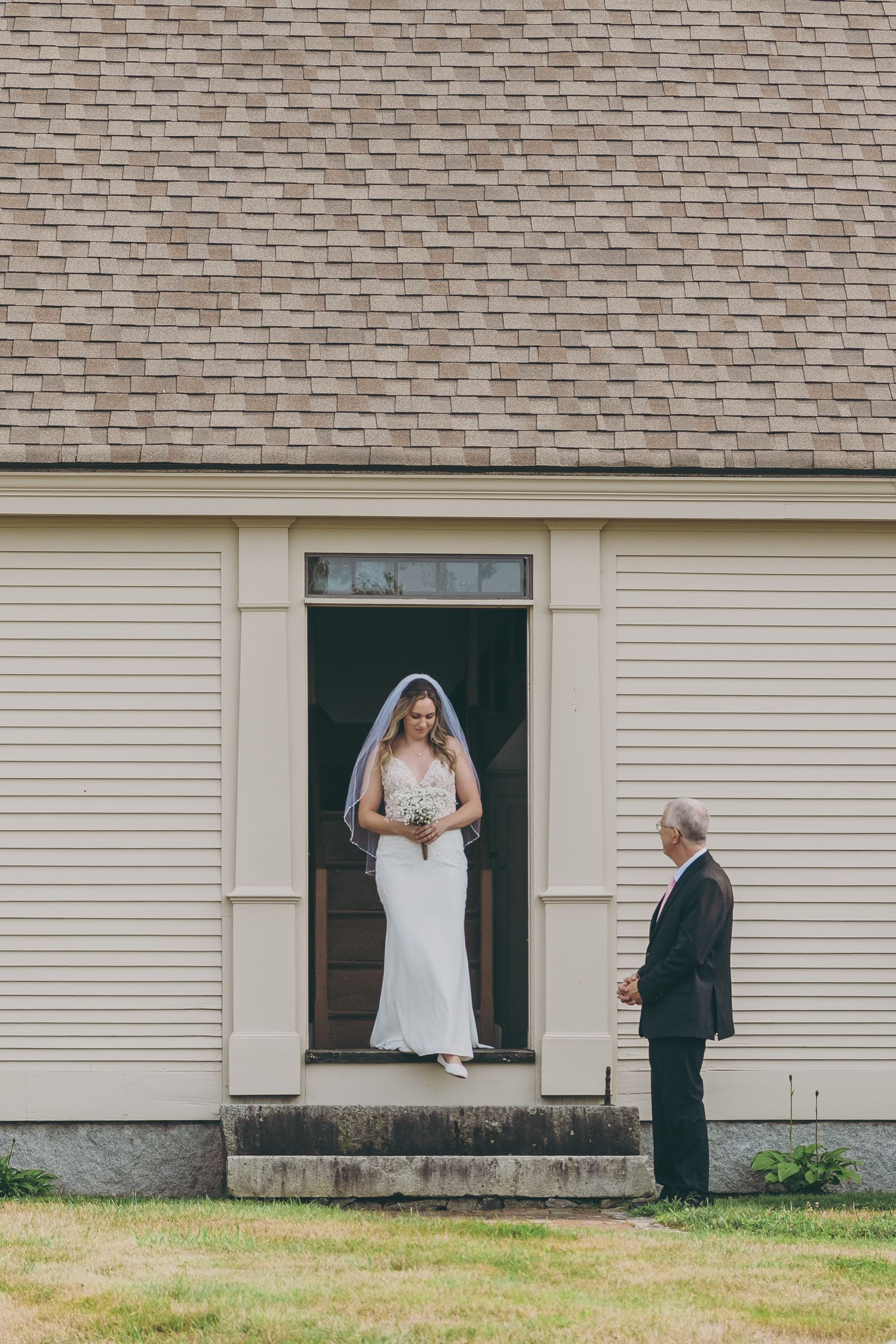 Bride in white gown stands in doorway while attendant waits outside beige building.