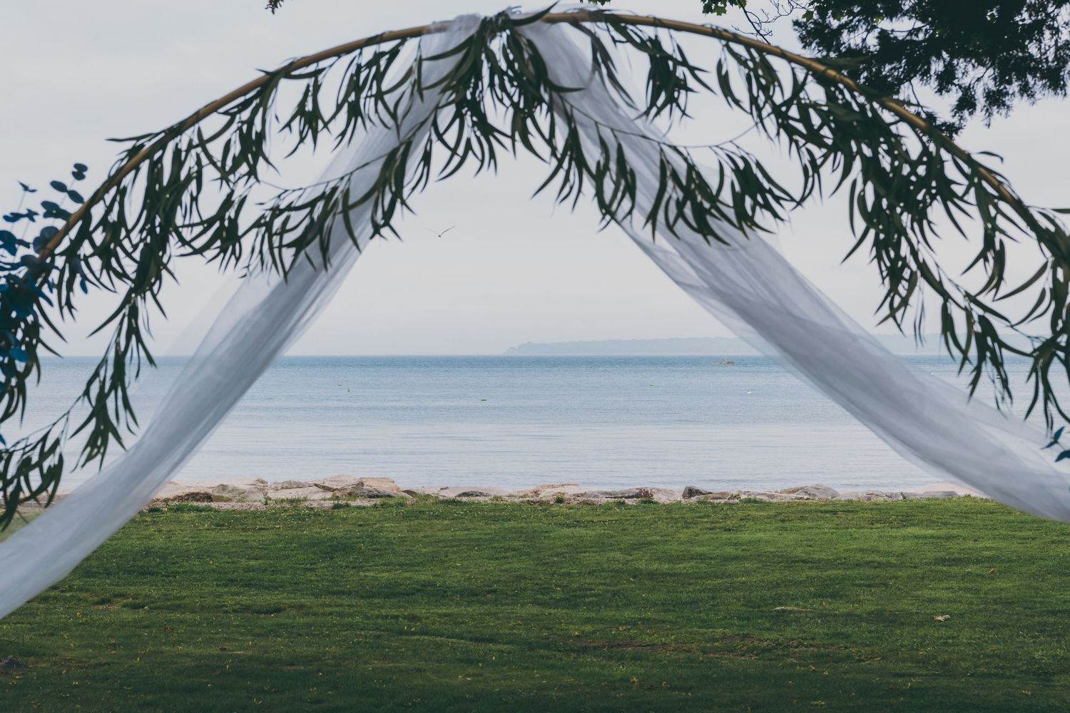 White wedding arch draped with greenery frames a serene ocean view across a manicured lawn setting.