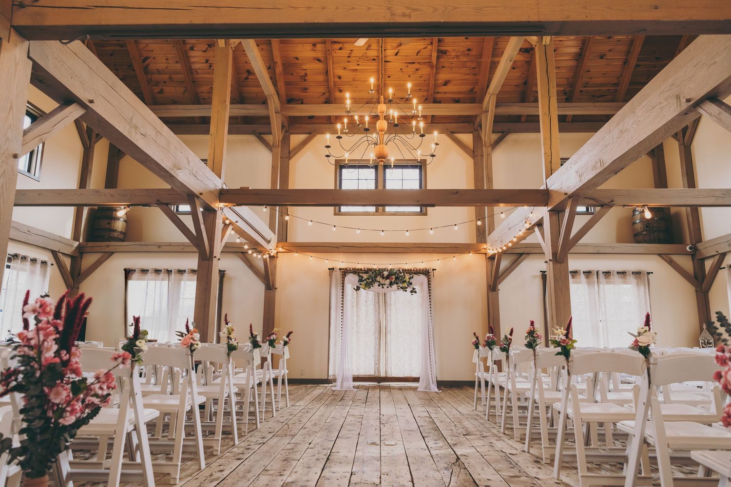 Rustic wooden beam barn venue decorated for wedding ceremony with white chairs and chandelier lighting.