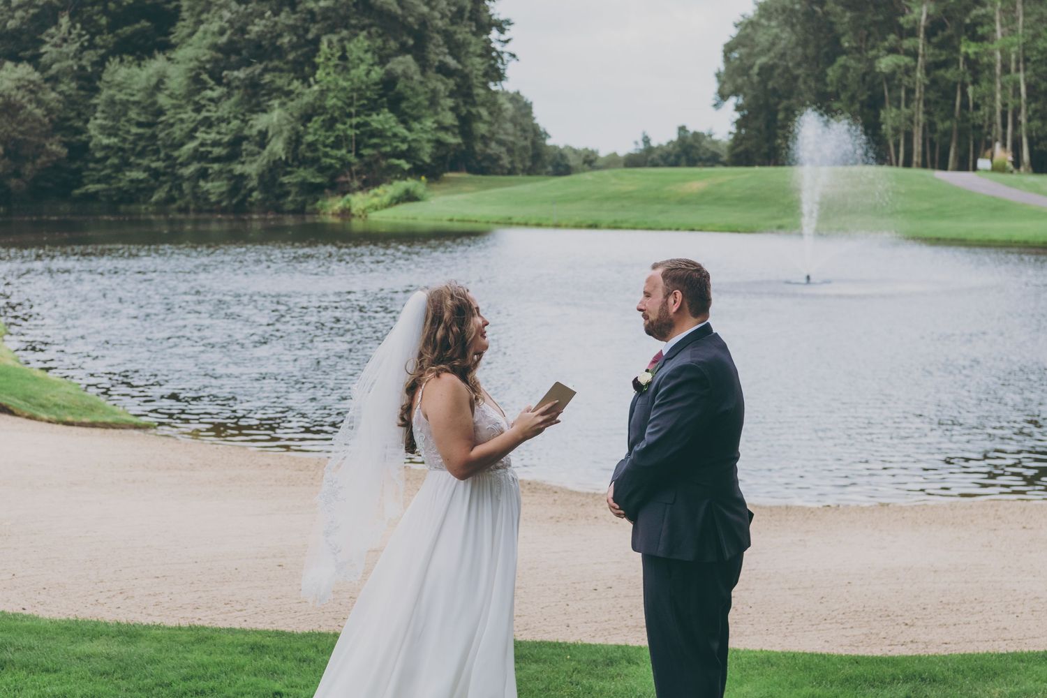 A scenic wedding ceremony by a pond with a water fountain in the background at an elegant outdoor venue.