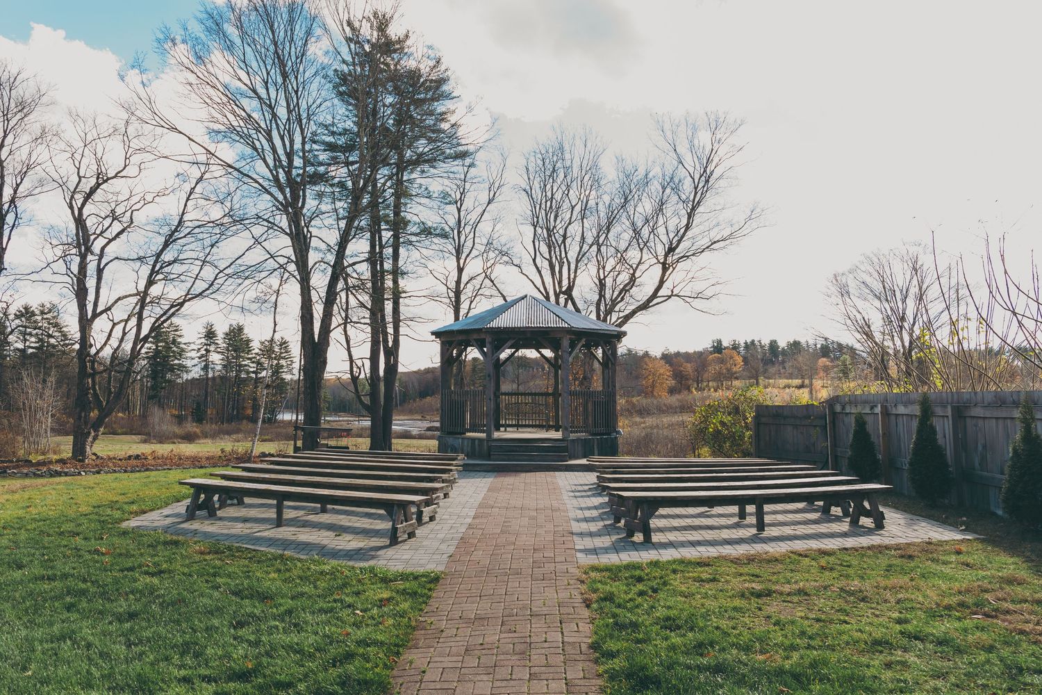 A rustic wooden gazebo with picnic benches on a grassy lawn surrounded by bare winter trees.