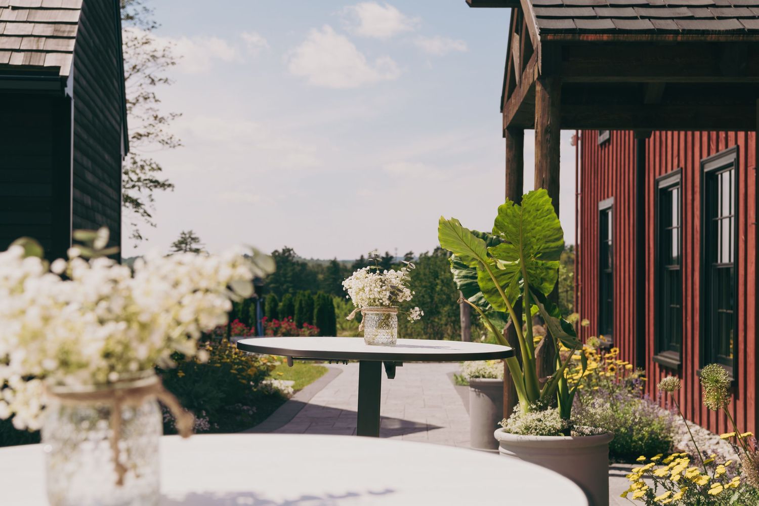 Outdoor wedding venue terrace with white floral arrangements and potted plants between rustic barn-style buildings.