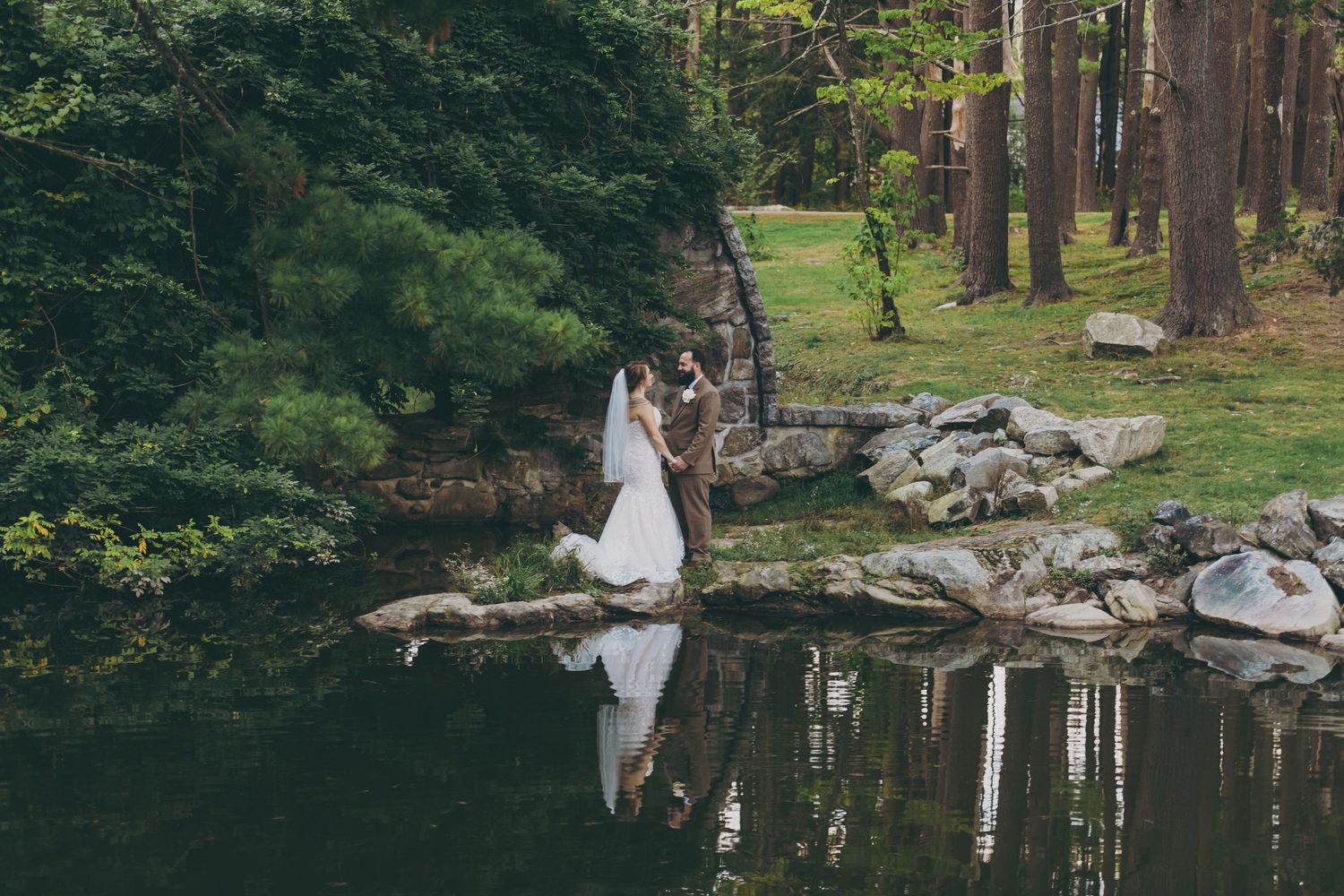 Wedding couple reflected in tranquil pond surrounded by forest landscape and rocks.