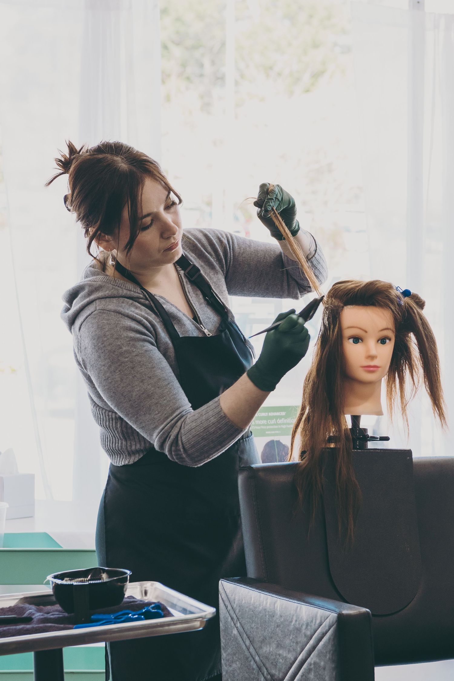 Stylist in grey sweater and black apron practicing hair techniques on a mannequin head.