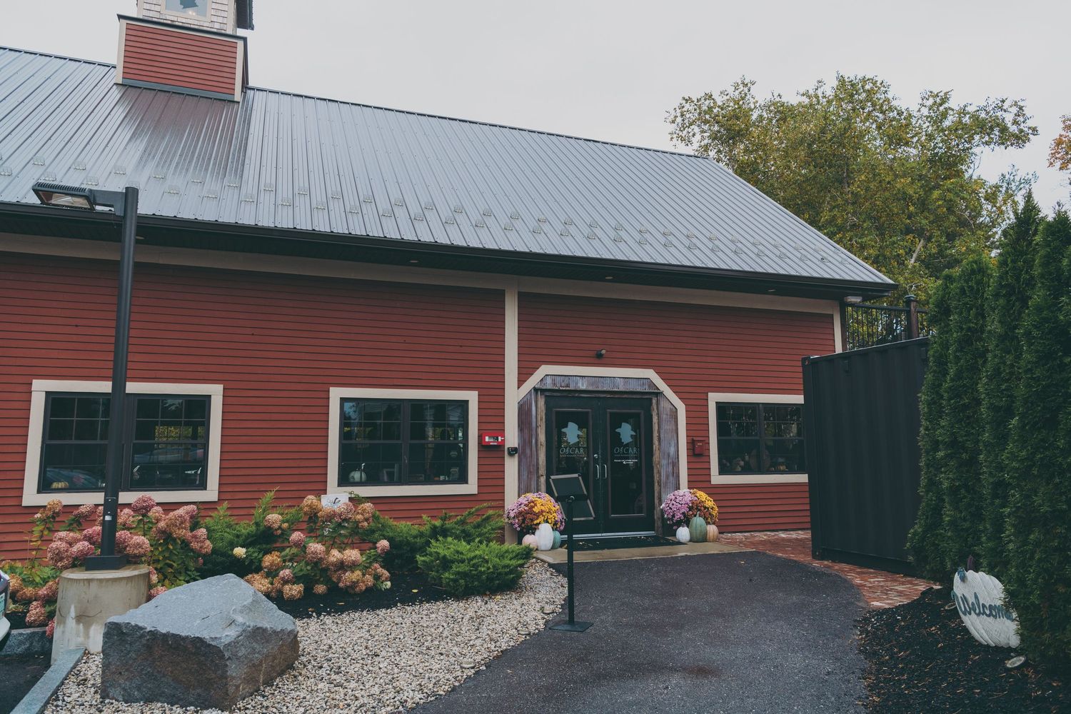 Red barn and storage buildings with metal roofs and paved driveway entrance in a rural setting.