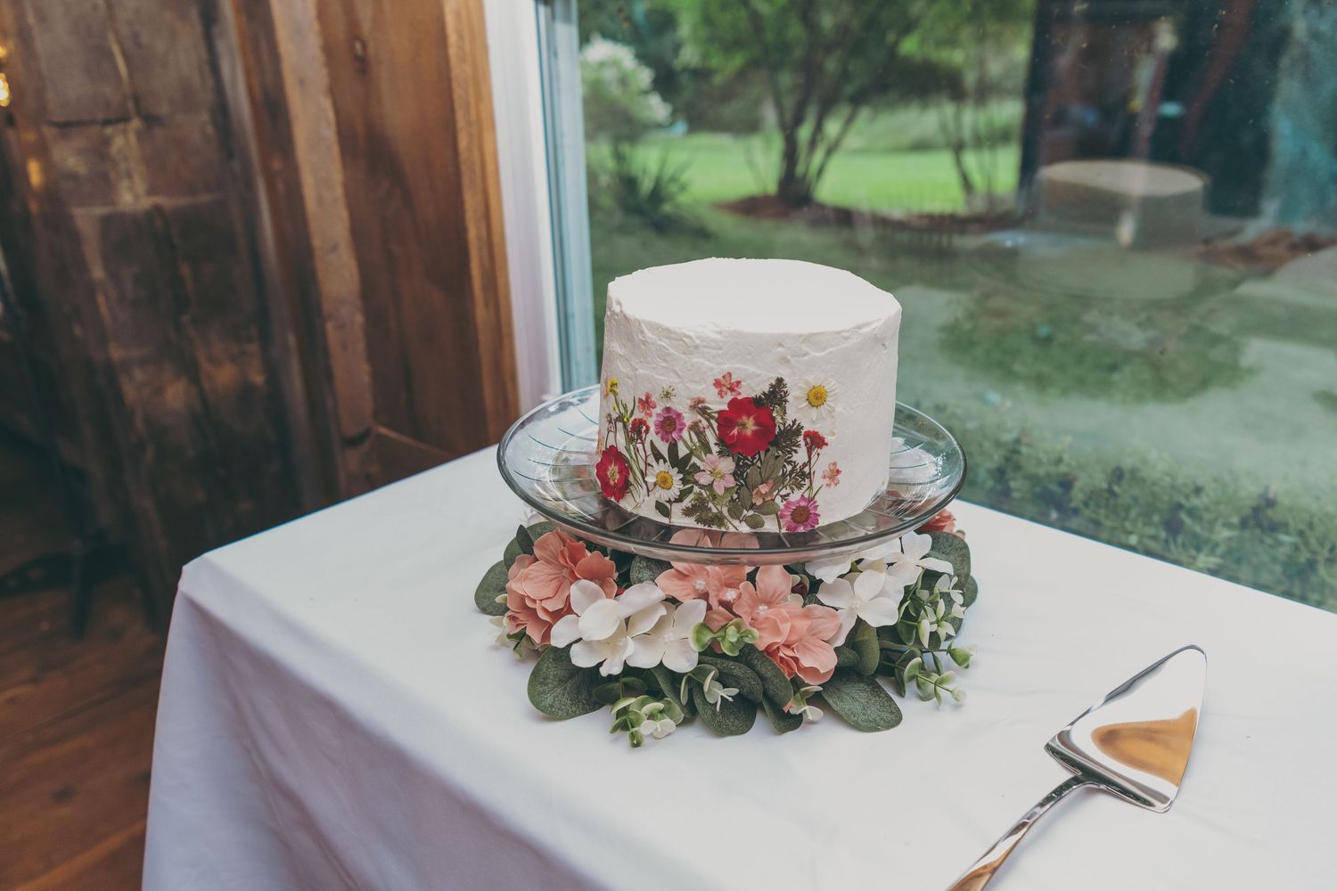 Wedding cake decorated with pink and red floral details sits on white tablecloth next to window with garden view.