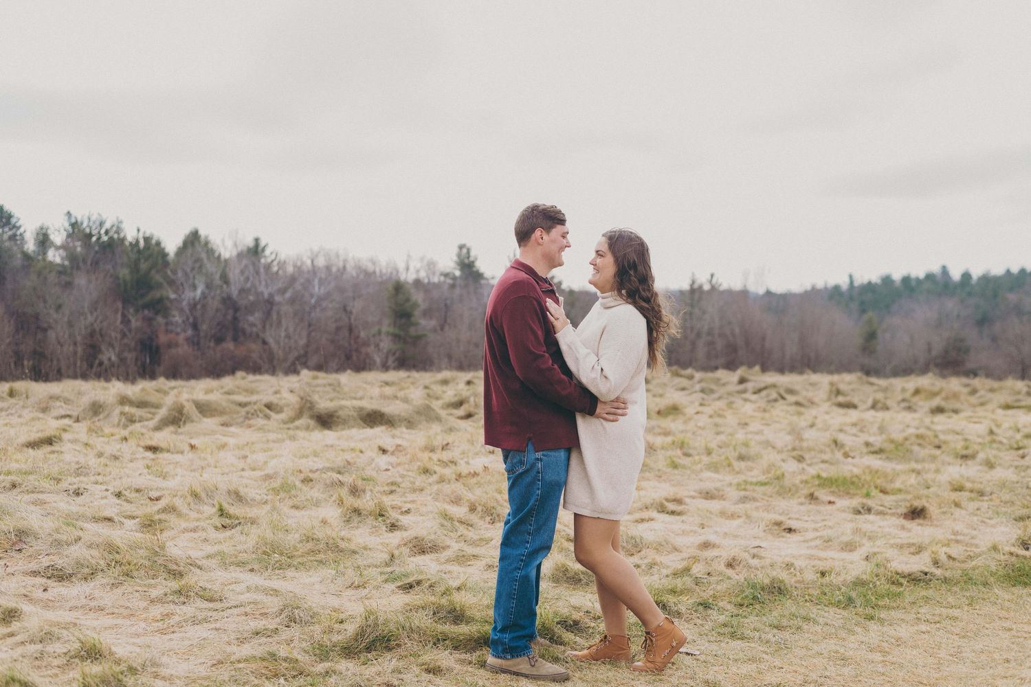 Couple in casual attire embrace in an open field with trees in the background on an overcast day.