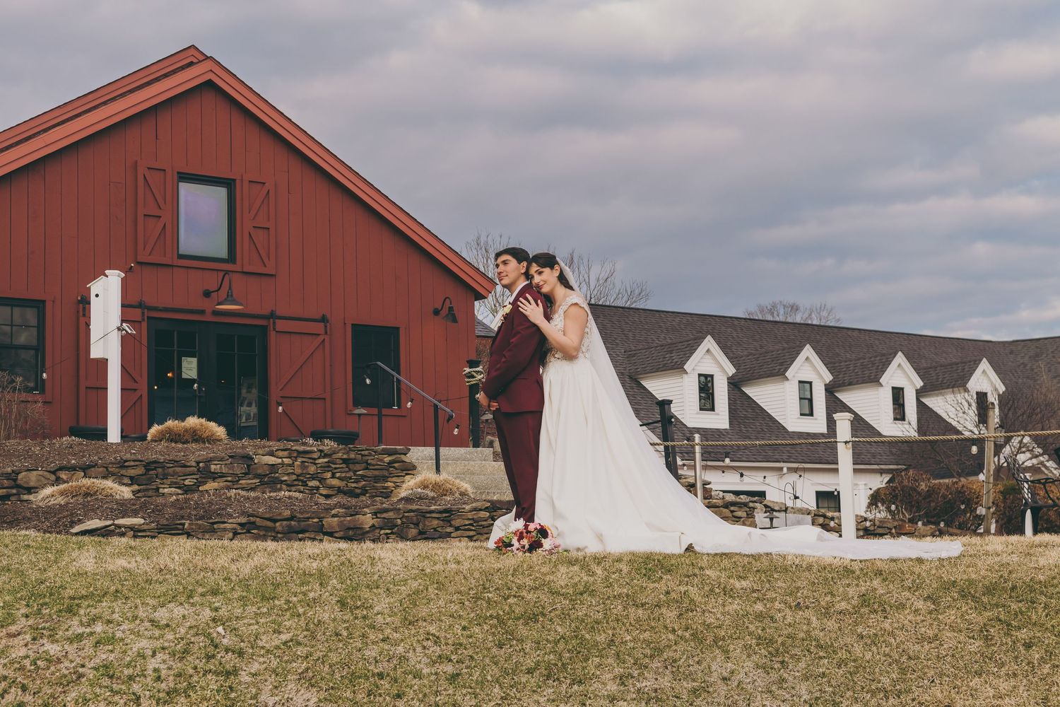 Wedding couple poses together on scenic hillside with rustic red barn and white buildings in background.