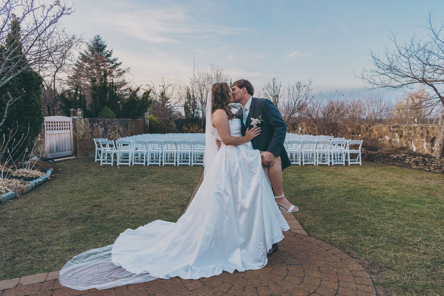 A wedding couple shares an intimate moment on a brick patio with white ceremony chairs in the background.