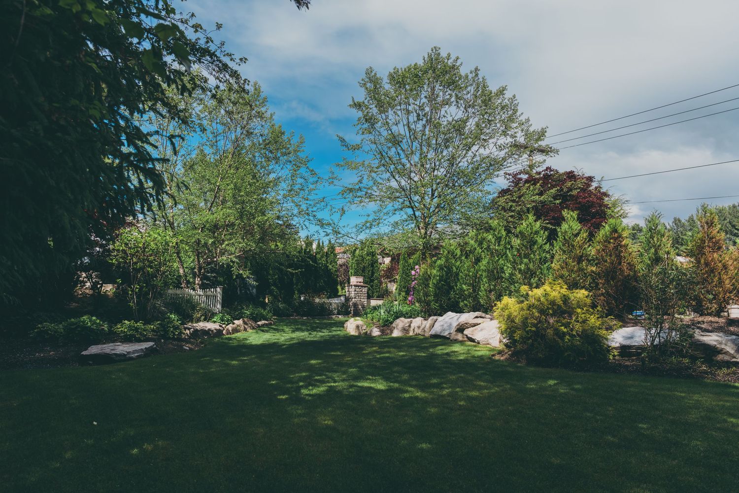 A landscaped backyard garden features green lawn, stone walls, and mature trees under a blue summer sky.