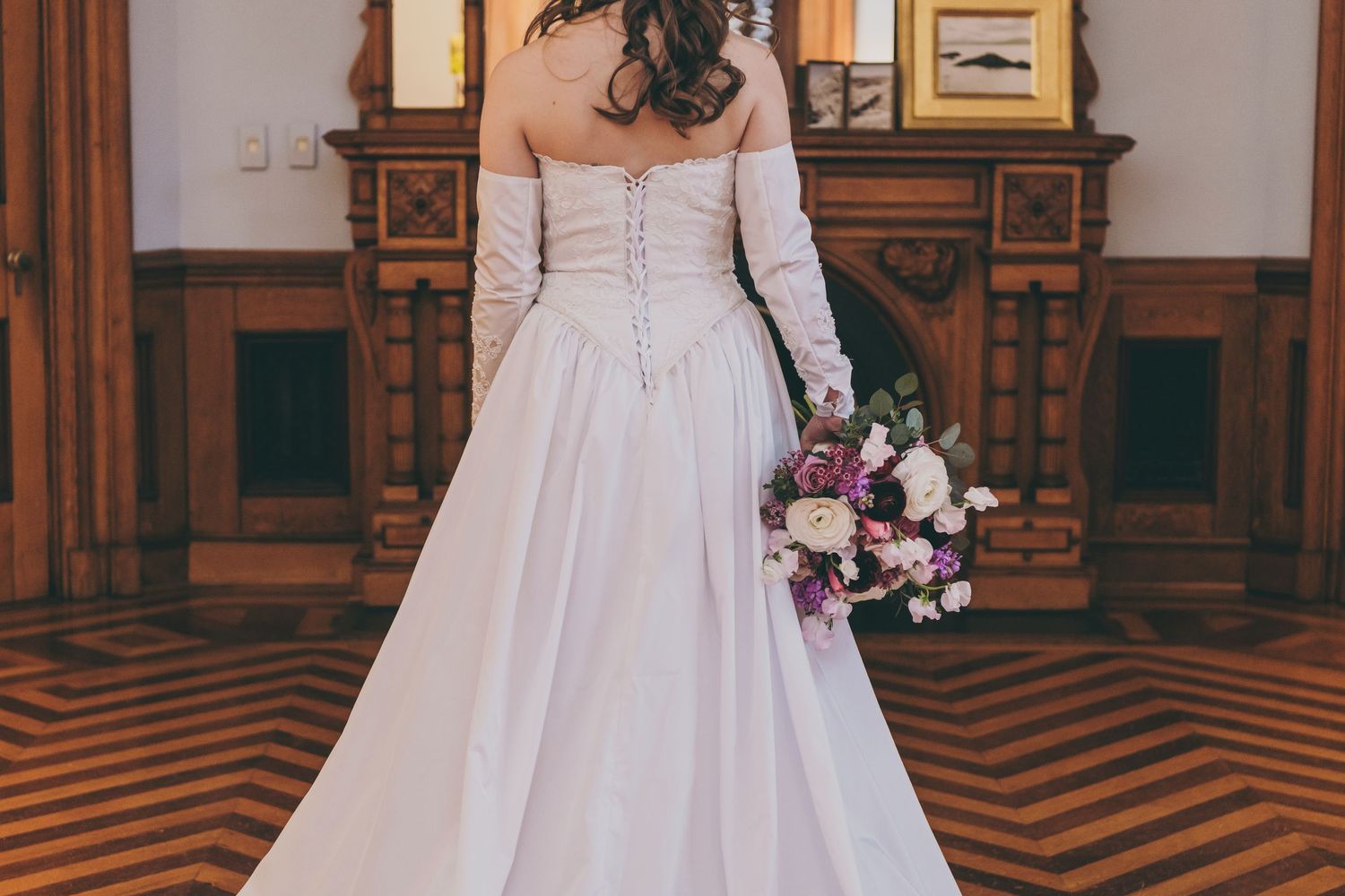Back view of a bride in an off-shoulder white gown with a floral bouquet against a chevron-patterned floor.