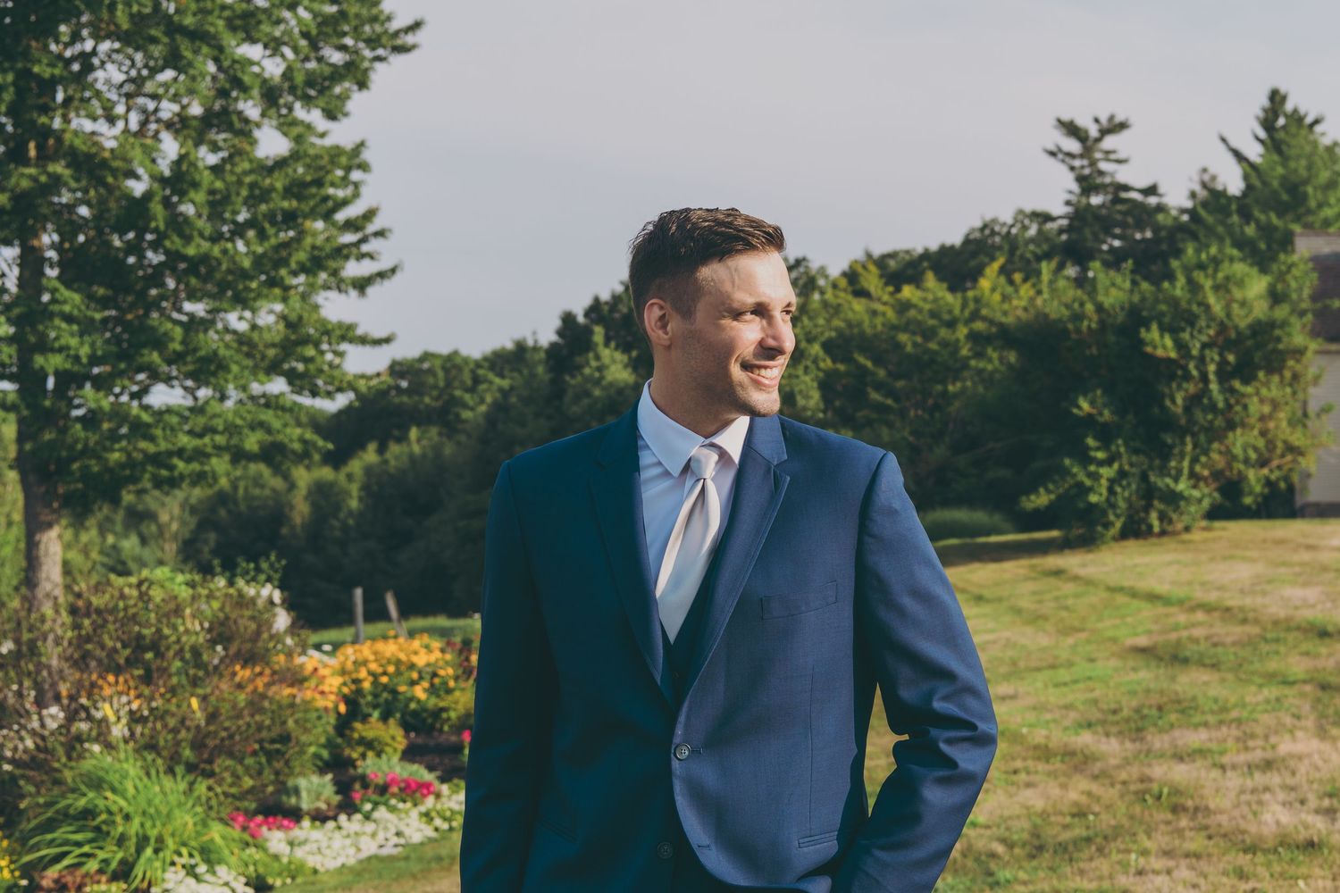 Groom in navy suit smiles while standing in garden with blooming flowers and trees.