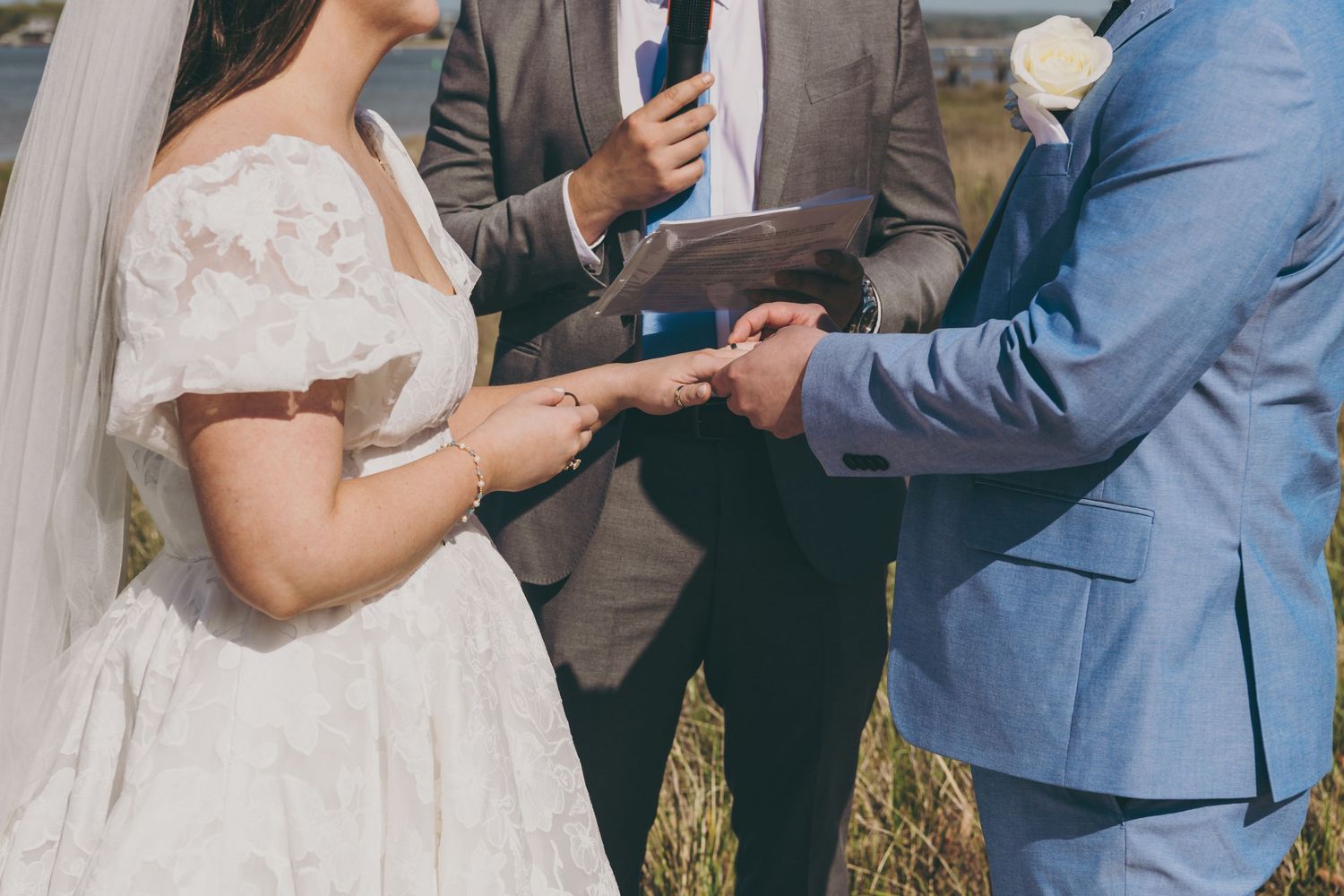 A couple exchanges rings during an outdoor wedding ceremony wearing traditional wedding attire.