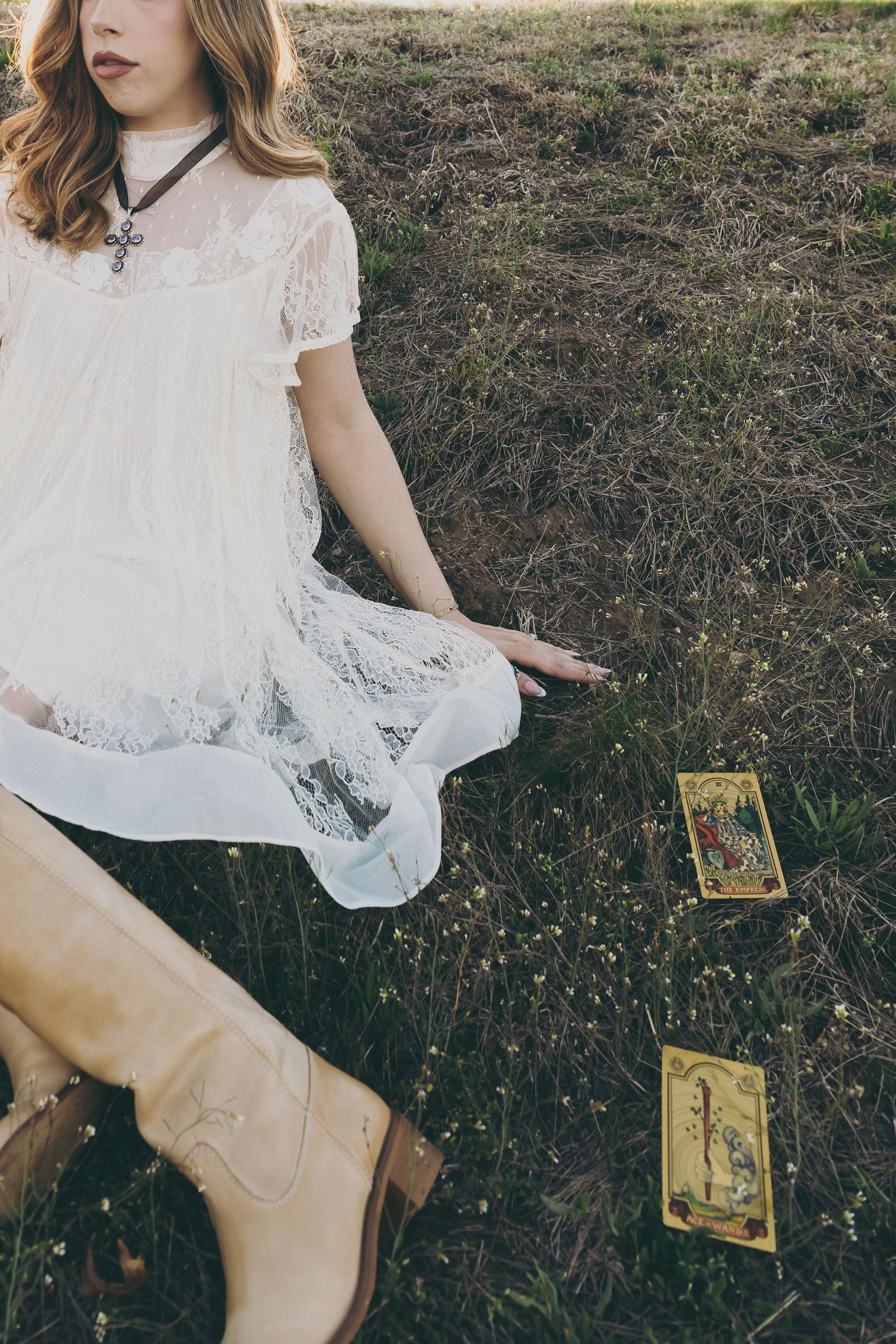 Person in white lace dress sitting on grass with tarot cards and brown boots.