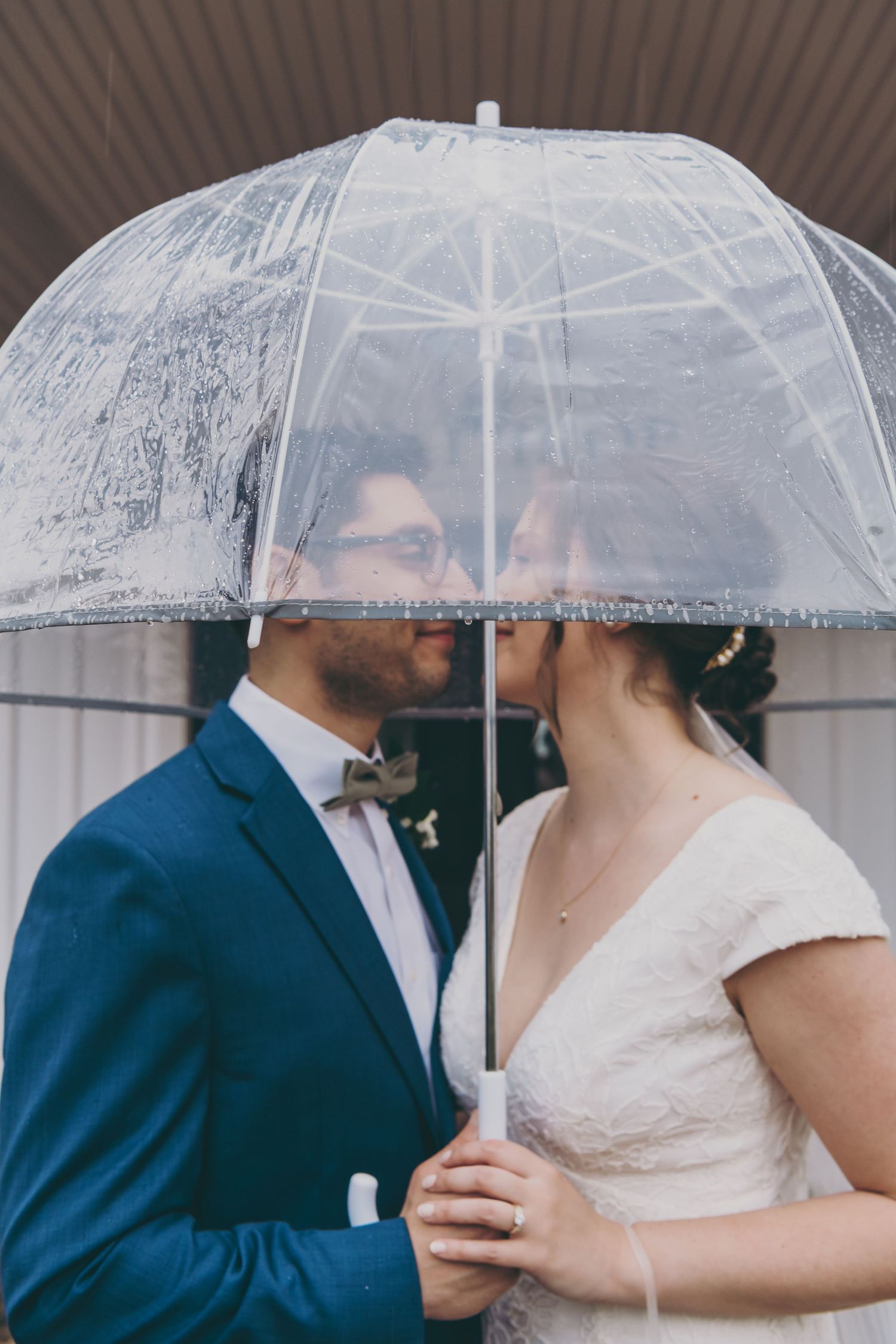 Wedding couple sharing intimate moment under clear umbrella in romantic rainy day portrait.