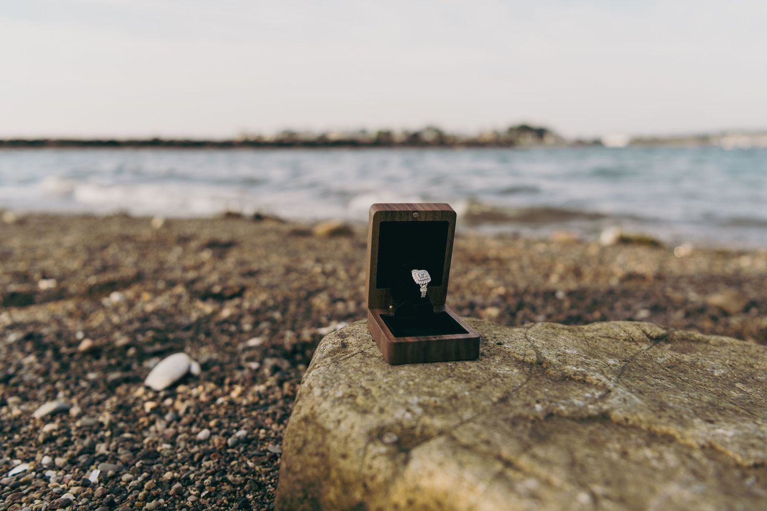Engagement ring in black box sitting on rocky beach coastline with ocean waves in background.