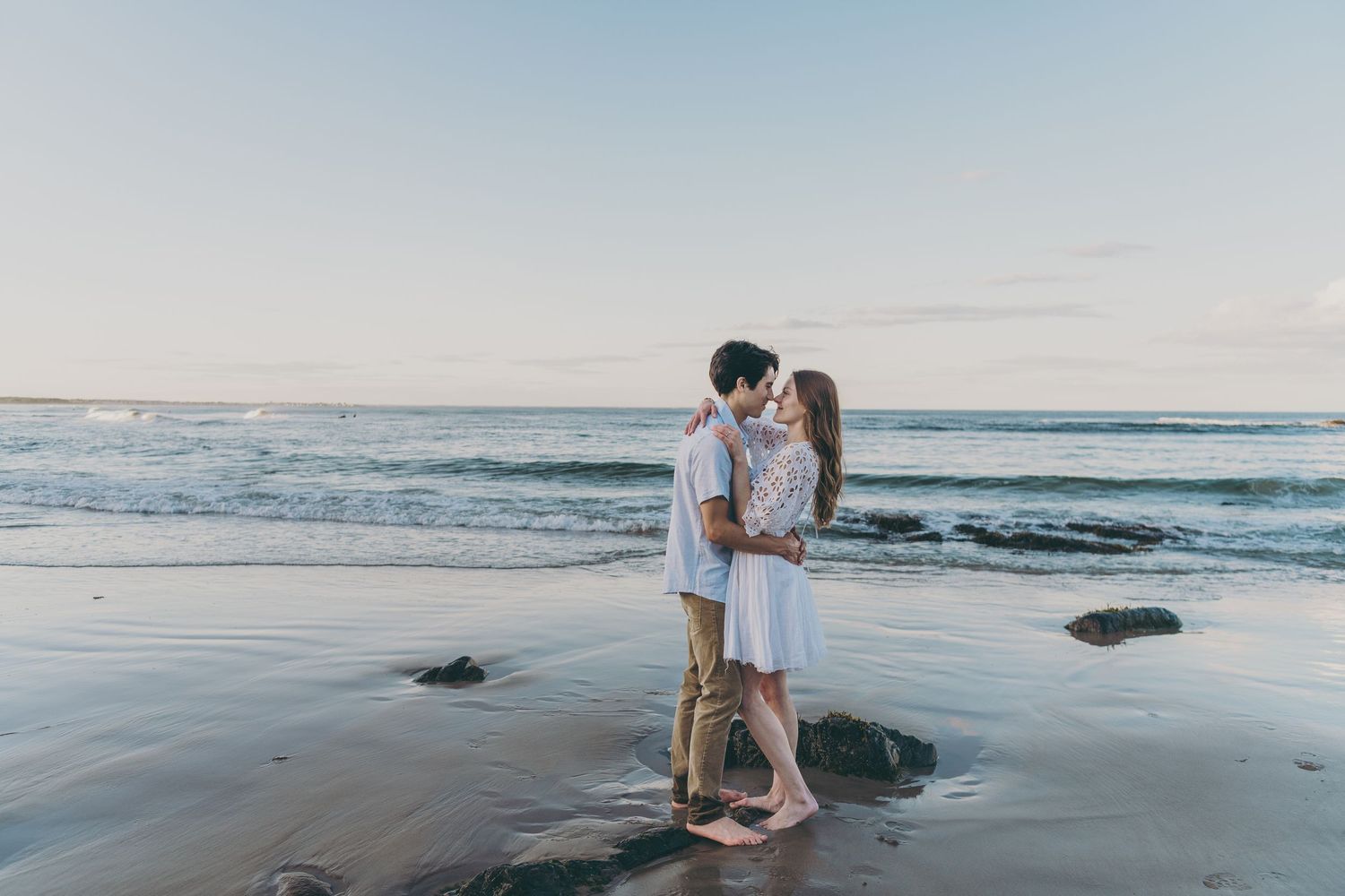 A romantic couple embraces on sandy beach with ocean waves and sunset sky behind them.