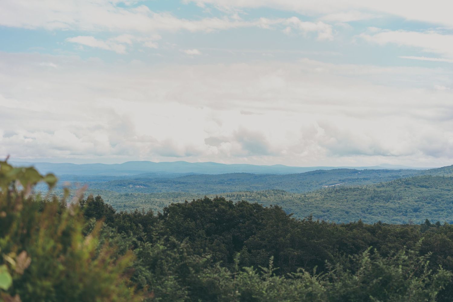 Scenic aerial view of rolling green hills and forests stretching to the horizon under a cloudy blue sky.