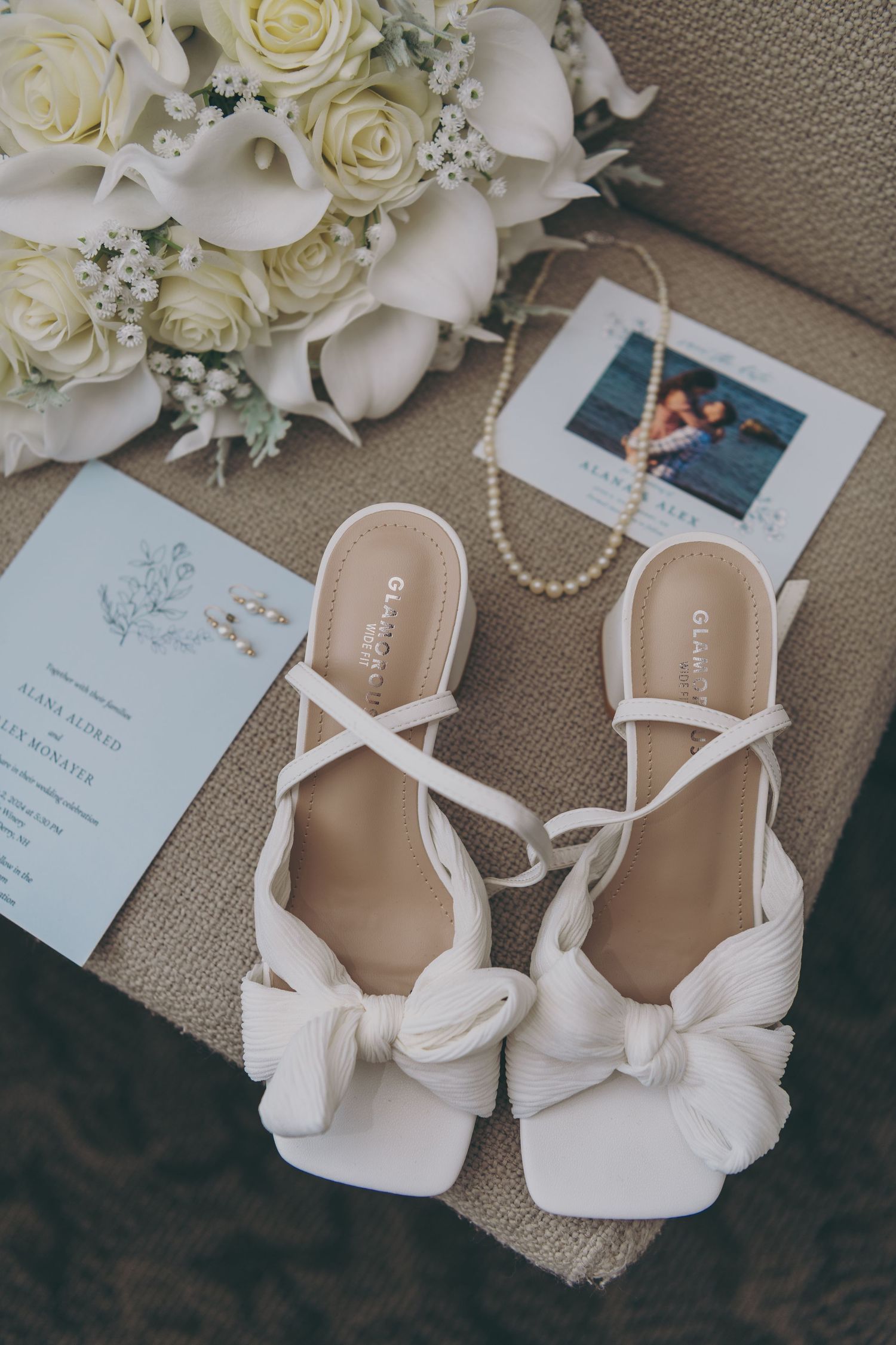 Elegant white bridal heels with pearls and roses arranged beside wedding stationery on a beige surface.